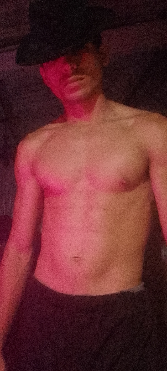 A grainy, dimly lit, vertical photograph depicts a single adult male subject from the mid-chest upward. The specific location in Juan Griego, Venezuela, is not discernible from the visual evidence. The subject wears a dark, wide-brimmed hat tilted forward, partially obscuring the upper portion of the face, though a mustache is visible on the upper lip. The upper torso is bare, revealing a lean to athletic physique with visible muscle definition in the chest and shoulders, and a visible navel. Dark-colored bottoms are visible at the waistline. The scene is illuminated by a prominent red or pink light source originating from the viewer's left, casting a strong magenta glow across the left side of the subject's face, neck, left arm, and torso. The right side of the subject and the background are rendered in deep shadow, appearing dark brown to black and featureless. The subject is static, appearing to pose for the photograph. No other objects, individuals, or interactions are discernible within the frame.