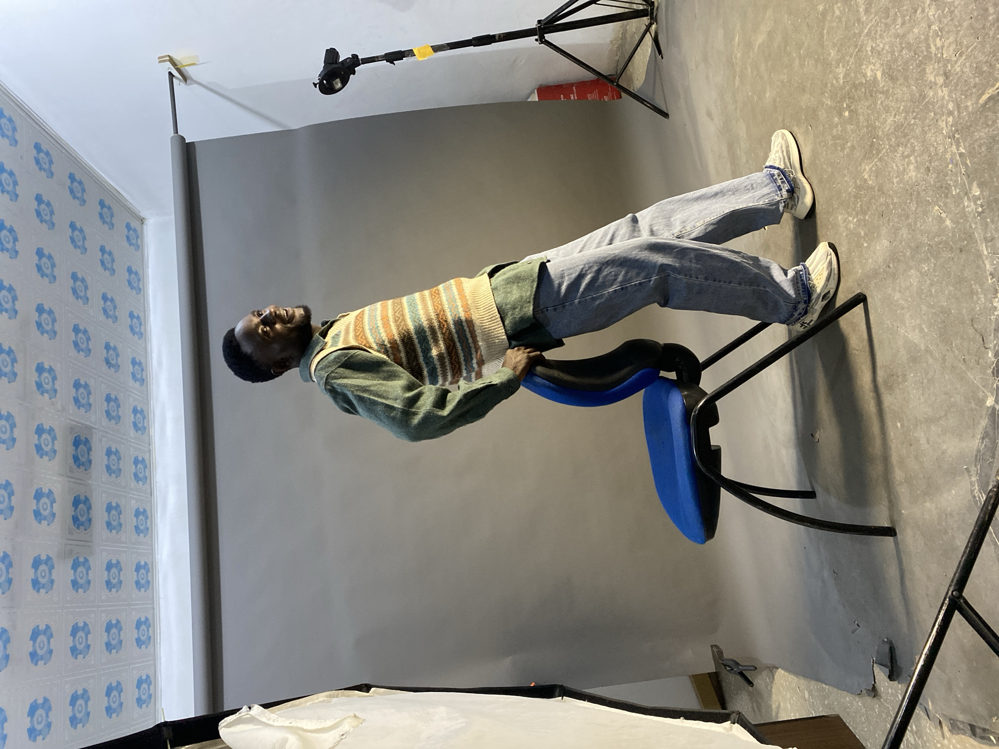 The image depicts a man reclining on a blue office chair in a photography studio. He is wearing a green shirt, a striped sweater vest, and blue jeans. He is facing toward a wall with a blue patterned wallpaper. Behind him is a grey backdrop. Lighting equipment, including a light stand and diffusion panel, are visible. The location is implied to be Jos, Nigeria, based on the prompt.