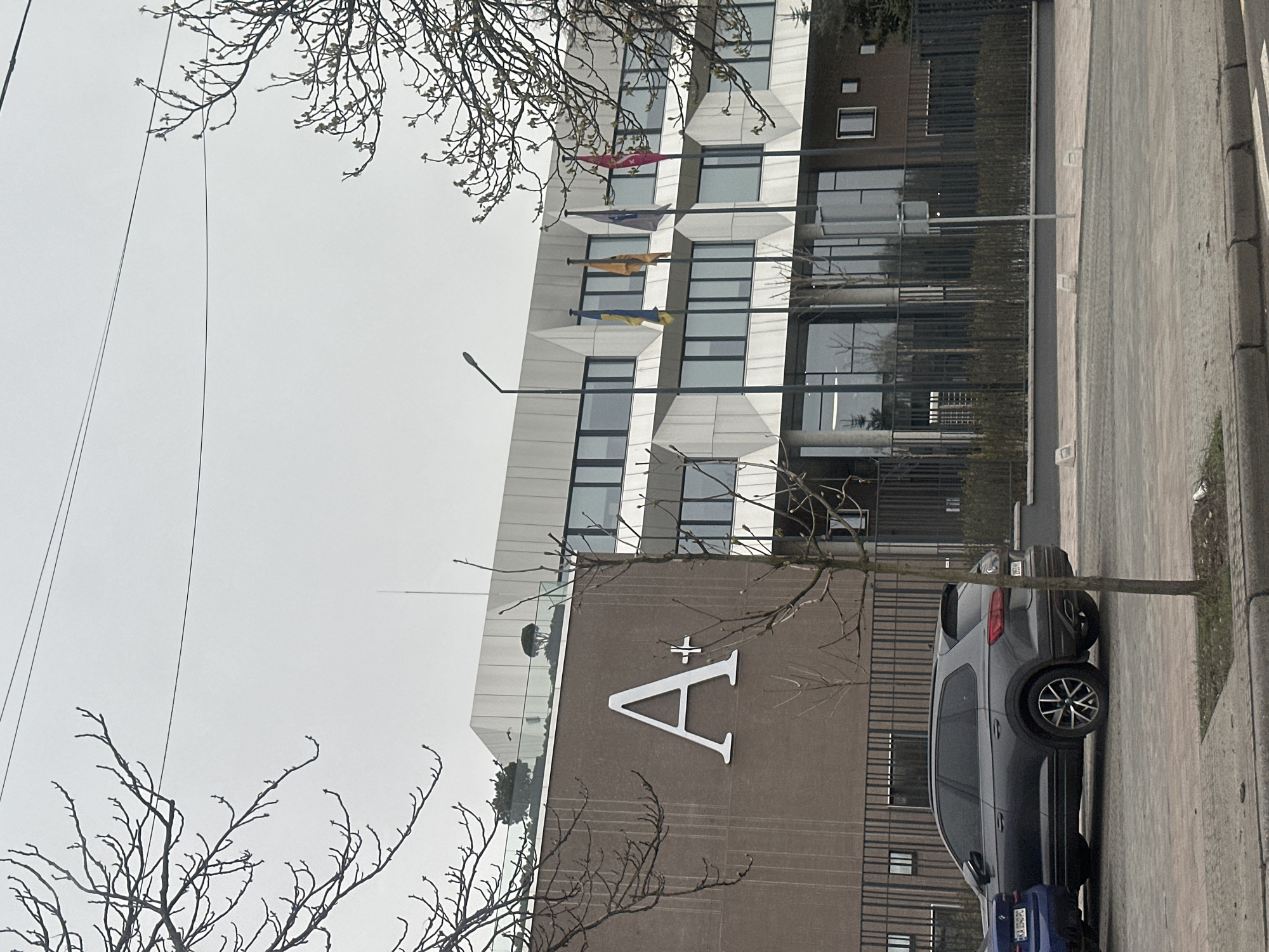 The image shows a building in Kyiv, Ukraine. It is a modern building with a large "A+" logo on the facade. Several flags hang from the building. A car is parked on the street in front of the building. The sky is overcast, and trees are visible in the foreground.