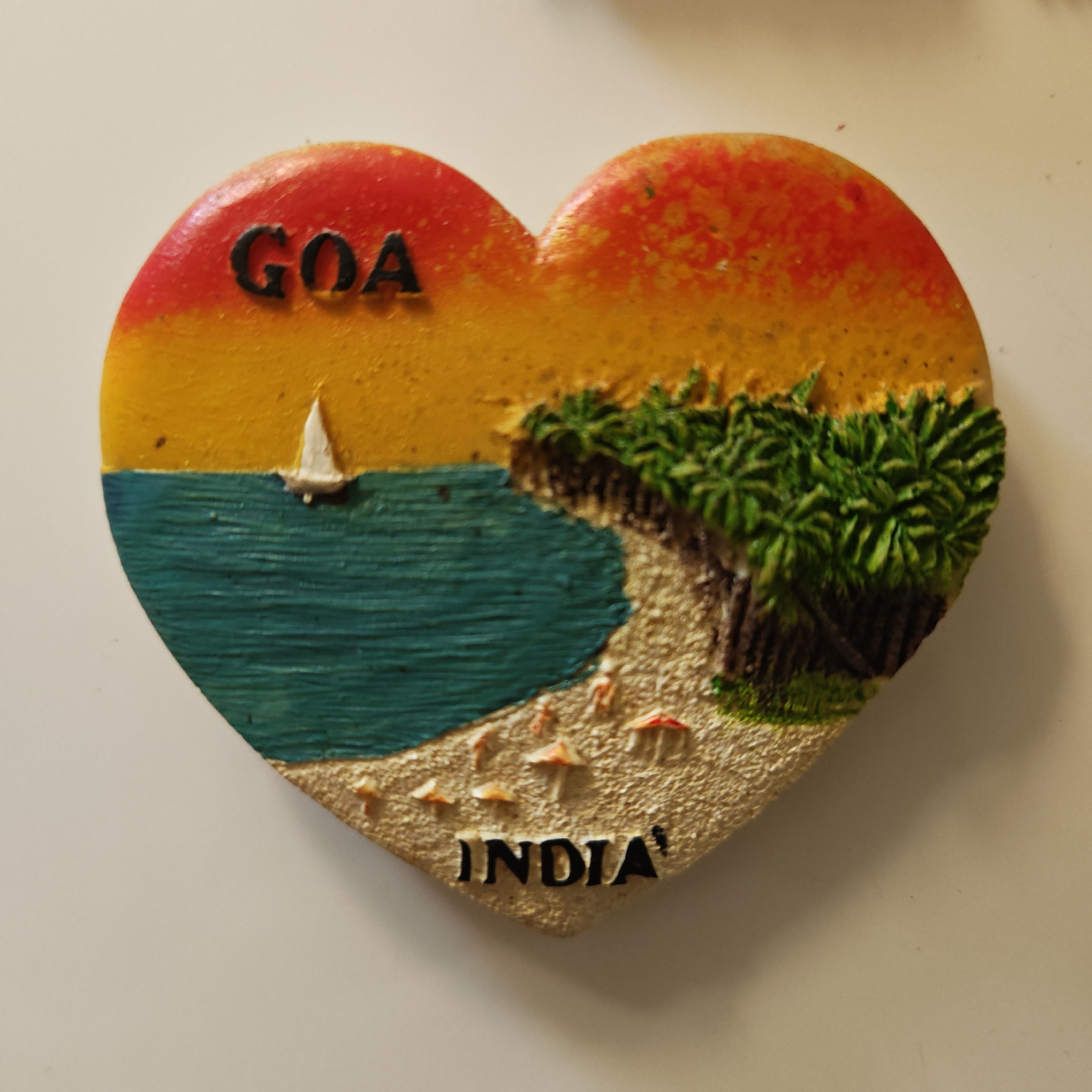 A heart-shaped fridge magnet featuring a vibrant, three-dimensional beach scene is depicted in the image. The magnet is predominantly colored with a gradient sky, transitioning from red at the very top to orange and yellow across the upper half, suggesting either a sunrise or sunset. Below the sky, a body of turquoise blue water with visible ripples represents the sea or ocean. A small white sailboat with a triangular sail is visible on the water, positioned towards the left side of the magnet.
The right side of the magnet portrays a sandy beach adorned with multiple lush green palm trees, indicating a tropical setting. On the sandy shore, several small, stylized structures with red caps and white stems are scattered, possibly representing beach umbrellas or small mushrooms.
The magnet has raised textures, giving depth to the palm trees, water, and sand. The word "GOA" is prominently displayed in black, uppercase letters on the upper left portion of the magnet, against the yellow sky. Towards the bottom, also in black, uppercase letters, is the word "INDIA". These texts clearly identify the depicted location as Goa, India. The overall scene evokes a sense of tropical vacation and serene coastal beauty. The background of the image is a plain, light-colored surface, likely a wall or refrigerator.