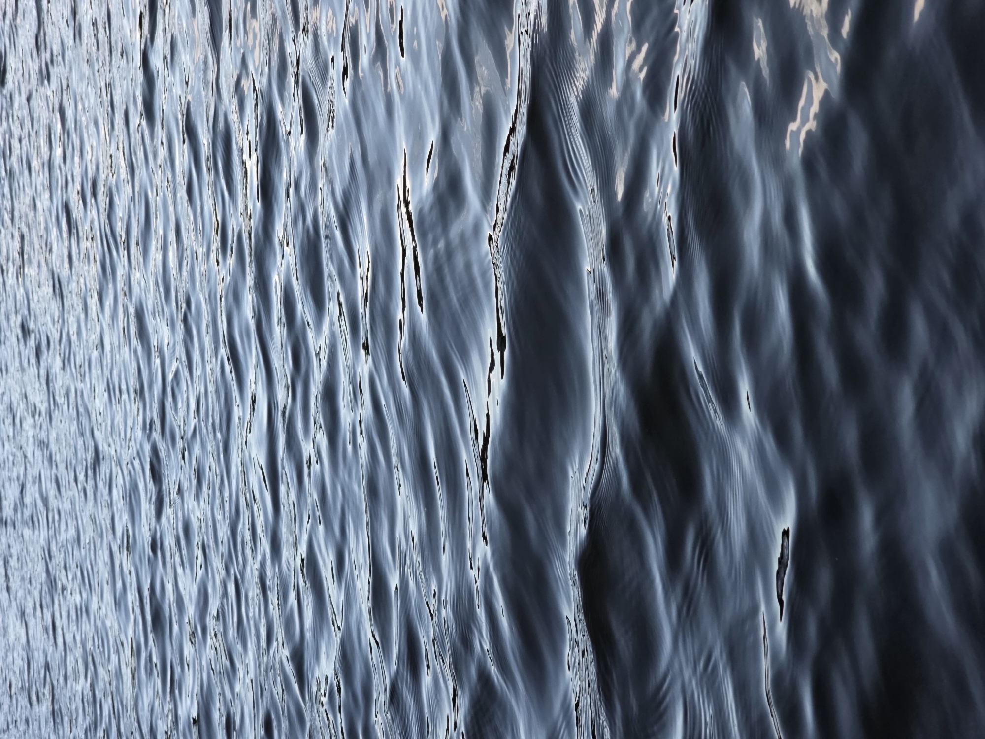 The image displays a close-up view of rippling water, likely a lake or river, with gentle waves creating a textured surface. The water is a deep blue-grey color, reflecting the sky or surrounding environment. There are highlights and shadows on the water's surface, emphasizing the movement and depth. The perspective is straight on, looking down at the water.
The location is the Ternopil Reservoir, also known as Ternopil Lake, in Ternopil, Ukraine. This reservoir is a prominent feature of the city, located within its urban area. The scene depicts a natural element of the city's landscape. There are no people, subjects, or objects other than the water itself visible in the image. The environment is a body of water under natural lighting conditions.