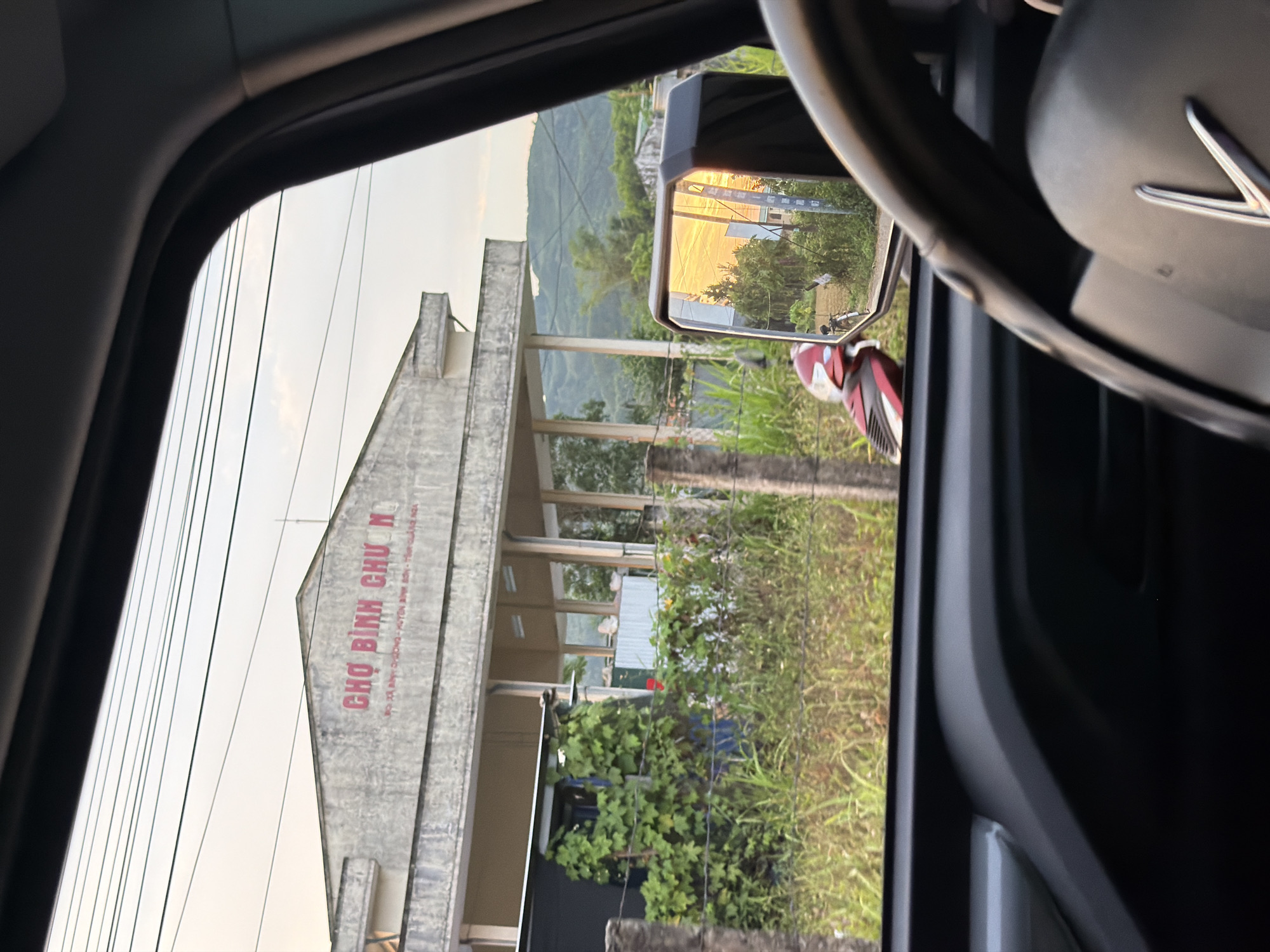 This media file captures an exterior scene in Bình Chương, Vietnam, viewed from the interior of a vehicle. The image is oriented vertically but depicts a landscape view from a car window.
On the left side of the frame, outside the vehicle, a concrete building with a prominent gable roof is visible. Large red text on the building clearly spells out "CHỢ BÌNH CHƯƠNG," which translates to "Bình Chương Market." Below this, smaller text further specifies: "ĐỊA ĐIỂM KINH DOANH CHỢ HỘ - XÃ BÌNH CHƯƠNG - HUYỆN BÌNH SƠN - TỈNH QUẢNG NGÃI," identifying it as a market business location within Bình Chương commune, Bình Sơn district, Quảng Ngãi province. Utility lines crisscross the sky above the building. Green foliage, including bushes and grass, grows in front of and around the building. In the background, rolling green hills or mountains are visible under a pale, soft sky.
To the right, elements of the vehicle's interior are seen, including the dark frame of the car window and a portion of a dark steering wheel. A side-view mirror captures a reflection of warm, golden light, possibly from a setting or rising sun, illuminating the road or landscape behind the vehicle. Partially visible below the mirror is a red and white object, possibly part of a scooter or motorcycle. The overall lighting suggests either early morning or late afternoon. The scene conveys a moment of observation while driving through or stopping near a local market in a rural or semi-urban Vietnamese setting.