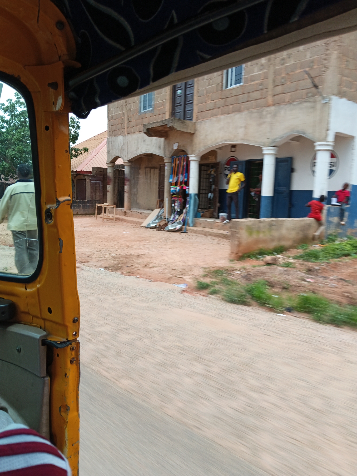 The image captures a street scene from the perspective of someone inside a yellow vehicle, likely an auto-rickshaw. In the foreground, the yellow door and part of the interior of the vehicle are visible, along with a person's shoulder wearing a striped shirt. The scene outside shows a dirt road and a building with a storefront.
Several people are present in front of the building. One man in a yellow shirt stands near the entrance, looking towards the right. Another person in a red shirt is partially visible, appearing to be sitting or leaning on a low concrete barrier. Further to the right, a person in a red shirt is walking away. The building has a facade with arches supported by pillars. Two large Pepsi logos are visible on the building's exterior, one on each side of the entrance.
The storefront appears to be selling various items, with racks of goods visible just inside the entrance. There is a small wooden table and some larger items, possibly metal basins, on the ground in front of the building. The ground is predominantly dirt and sand, with some patches of green grass and vegetation visible. The sky is overcast, suggesting it might be a cloudy day. The image appears to be taken during daylight hours. The overall impression is a bustling street scene in what appears to be a commercial area.