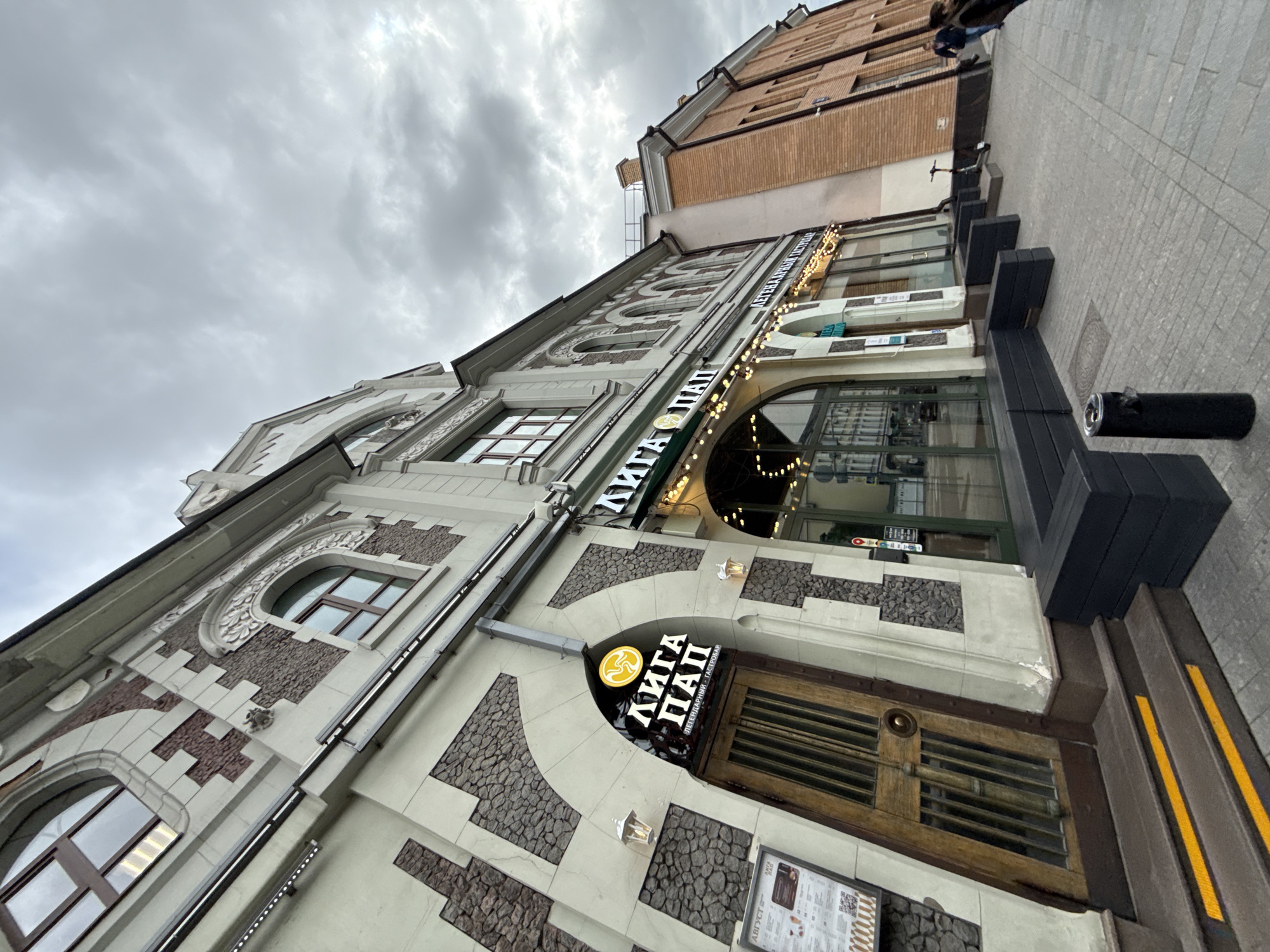 The image shows a view of a street in Moscow, Russia, on a cloudy day. In the foreground, there is a building with a decorative facade featuring stone accents and arched windows. A wooden door with metal grating is visible on the left side, with a sign above it displaying Cyrillic text and a circular logo. Next to the door is a menu board. The building to the right is made of red brick. The sidewalk is paved with gray tiles, and there are several black planters and a black trash bin on the right. A few people are walking in the distance, and an electric scooter is parked near the brick building. The overall scene depicts a commercial street with architectural details characteristic of the city.