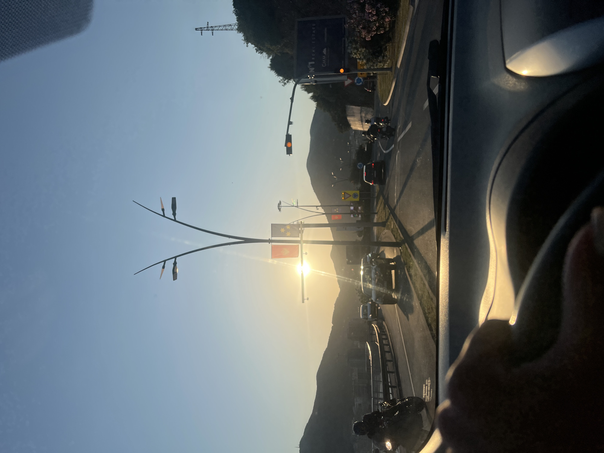 The image shows a road in Budva, Montenegro, as seen from inside a moving car. The sun is setting in the distance, partially obscured by mountains. Traffic is visible on the road, including cars and motorcycles. Flags of Montenegro and Budva are on poles along the road. A large billboard is also present. Traffic lights are visible and seem to be in operation. A power line tower is on the horizon. The steering wheel and part of the car's interior are visible in the foreground.