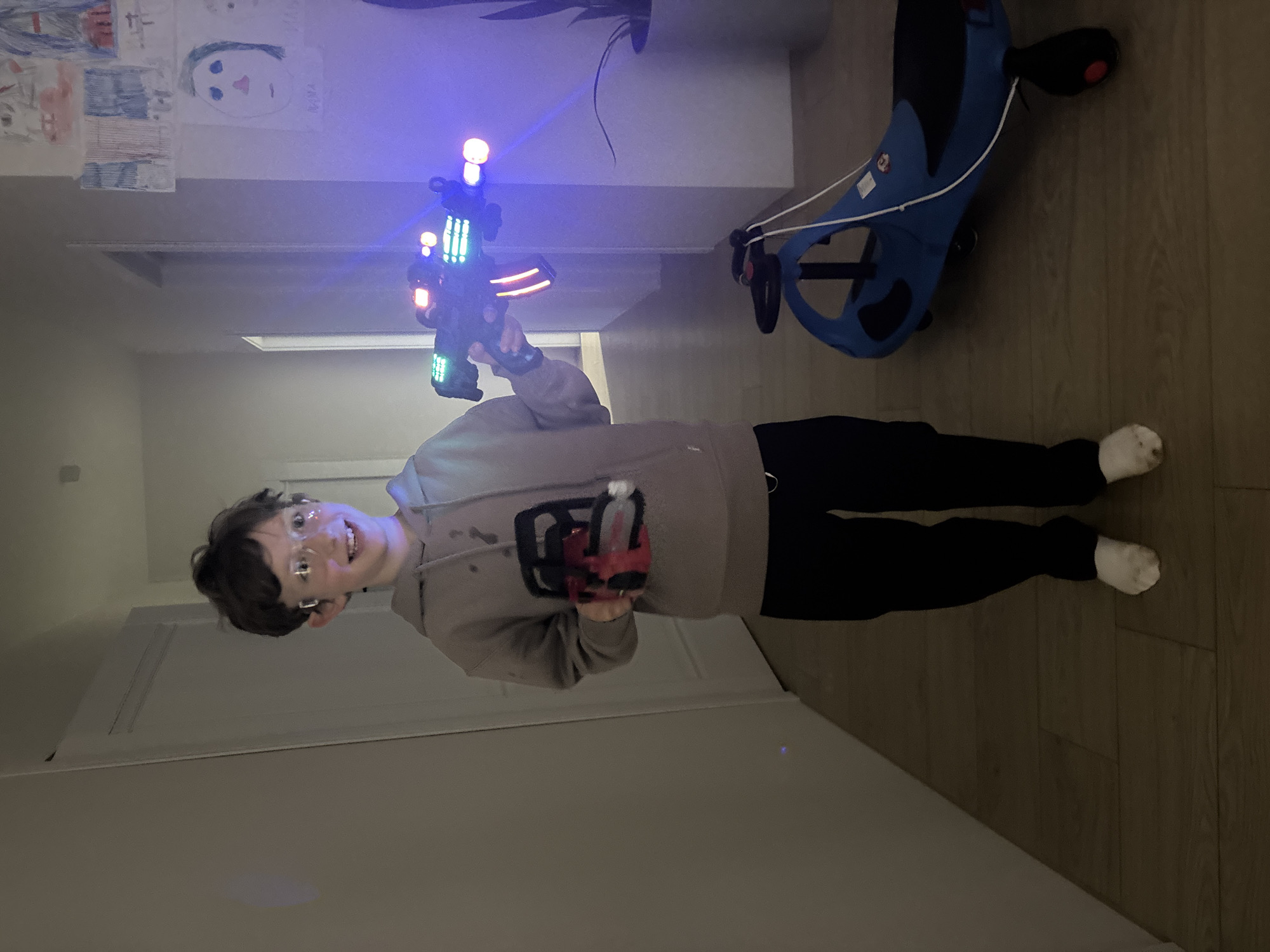A young person stands indoors, smiling and looking directly at the camera, holding two illuminated toy laser guns. The individual has short dark hair and wears glasses, a light grey hooded sweatshirt, dark trousers, and white socks. In their right hand, they hold a large, black toy gun that emits vibrant blue, green, and red lights. A smaller, red and black toy gun is held in their left hand, appearing to be attached to their arm.
The setting is a room with light-colored wooden flooring and neutral walls. On the wall behind the person, several children's drawings are displayed, including a simple portrait of a face. To the right of the person, a blue and black ride-on toy is visible on the floor. The lighting in the room is dim, accentuating the bright lights of the toy guns, which suggests it might be evening or nighttime. The person's happy expression indicates they are enjoying playing. This scene takes place in Chaikovsky, Russia. There is no clearly legible text visible in the image.
