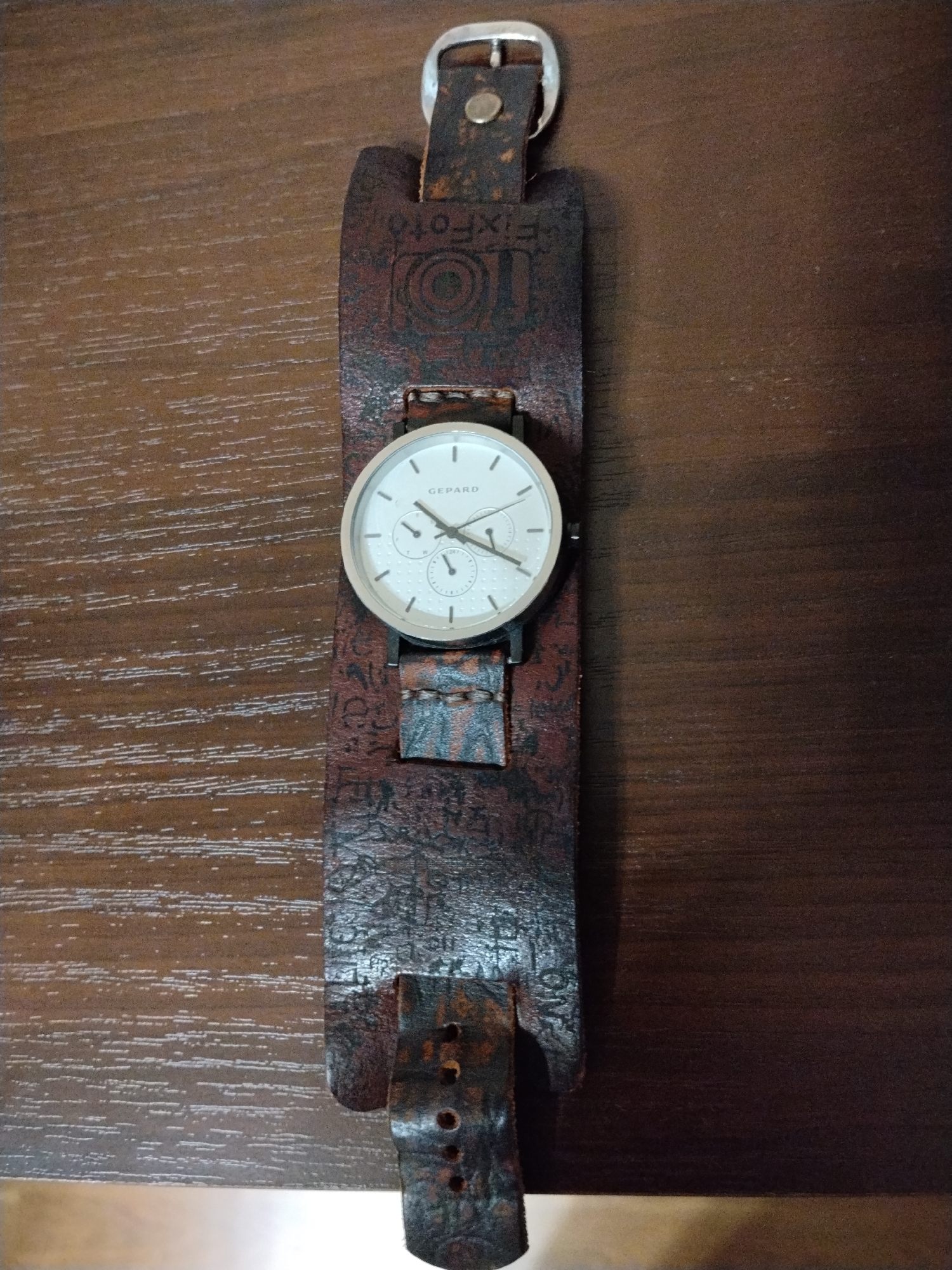 A Gepard brand wristwatch with a white face and silver-toned case is attached to a wide, brown leather cuff-style strap. The strap has a weathered appearance and features embossed text and designs, including what appears to be "FixFoto". The watch and strap are laid flat on a dark brown wooden surface with a visible grain pattern. The image is taken from a top-down perspective. There is no indication of any specific location in Krasnogorsk, Russia.
