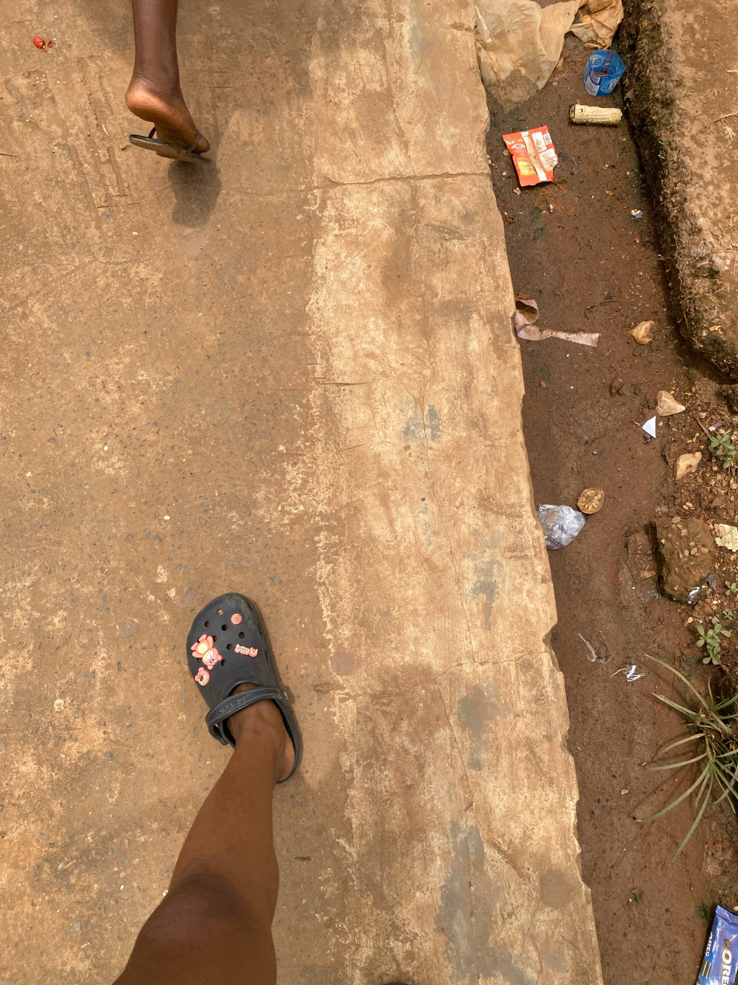 In Iworoko, Nigeria, a ground-level perspective reveals two individuals' feet. One person, wearing a flip-flop, is partially visible on the upper left, their heel slightly lifted. Below and to the left, another person's leg is prominent, adorned with a dark grey Croc shoe embellished with pink charms. The ground is a rough, concrete surface, appearing weathered and stained. To the right, a strip of muddy, uneven earth is strewn with various discarded items, including a red and white snack wrapper, a piece of corn cob, a crumpled plastic bag, and other unidentifiable debris. A small patch of green vegetation grows in the dirt. The scene suggests an outdoor urban environment, possibly a roadside or public walkway with a drainage ditch.