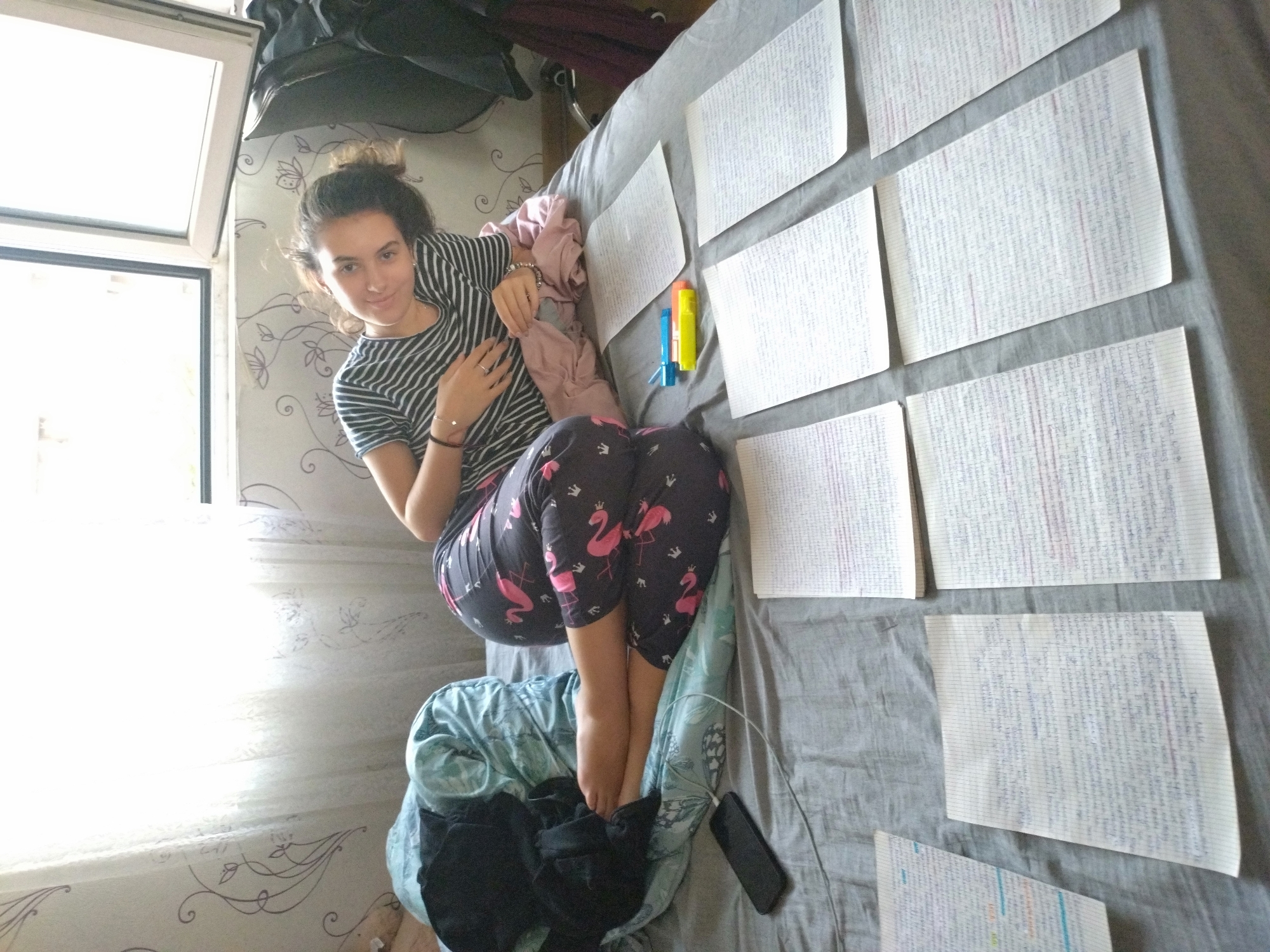 The image shows a young woman sitting on a bed. She has dark hair pulled into a bun and is wearing a striped t-shirt and flamingo-patterned leggings. The woman is looking at the camera and holding her hand to her chest. The bed is covered with a gray sheet and several pieces of paper covered in writing. There are also some colored markers and a smartphone on the bed. The background is a room with a window and patterned wallpaper. The location is likely in a private residence in the city of Stara Zagora, Bulgaria.