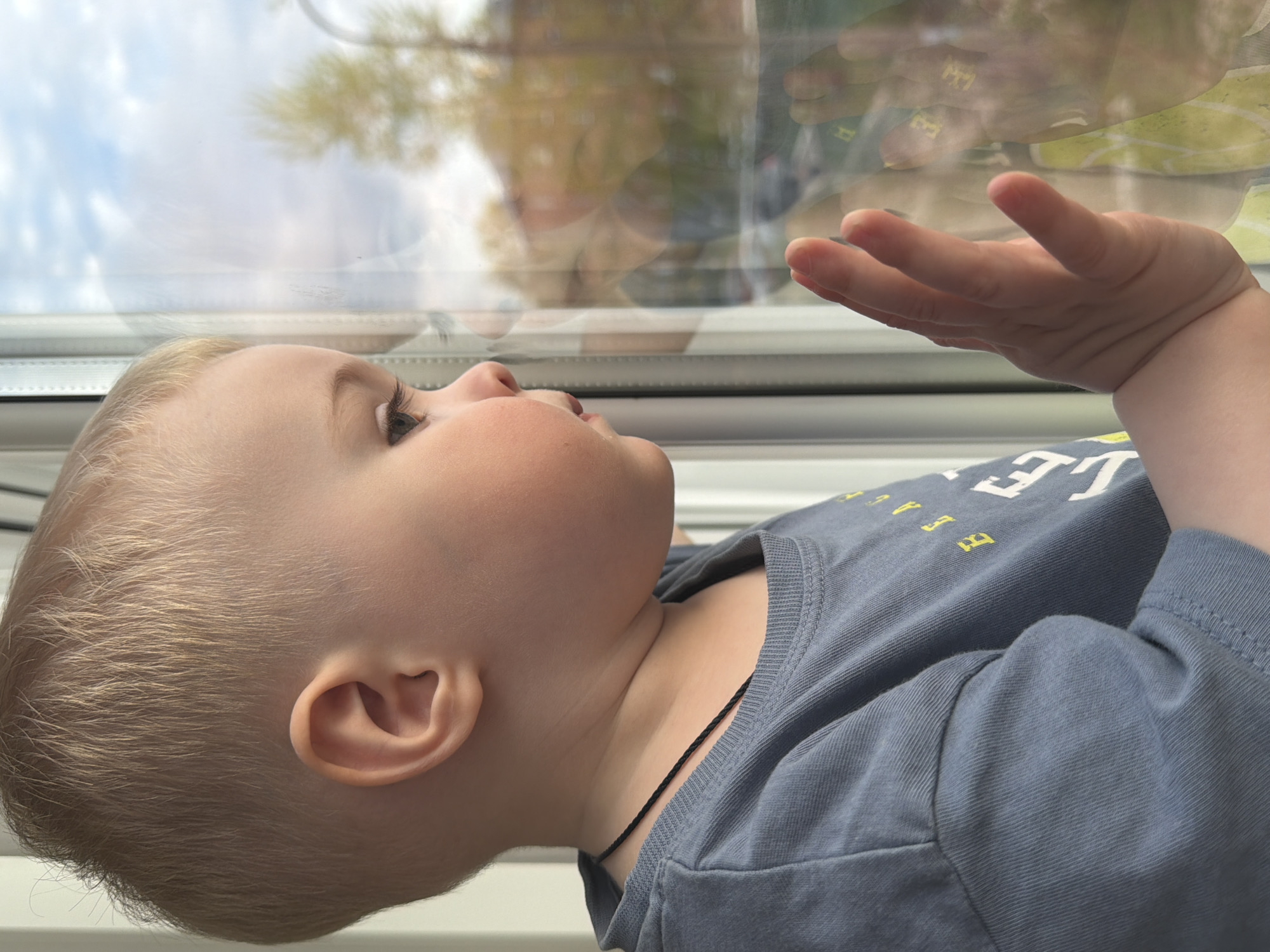 A blond-haired, light-skinned toddler is looking out a window, with their hand extended towards the glass. The child is wearing a blue t-shirt with a black cord around their neck. A reflection is visible in the window. The location is not identifiable as Votkinsk, Russia based on the visual information provided.
