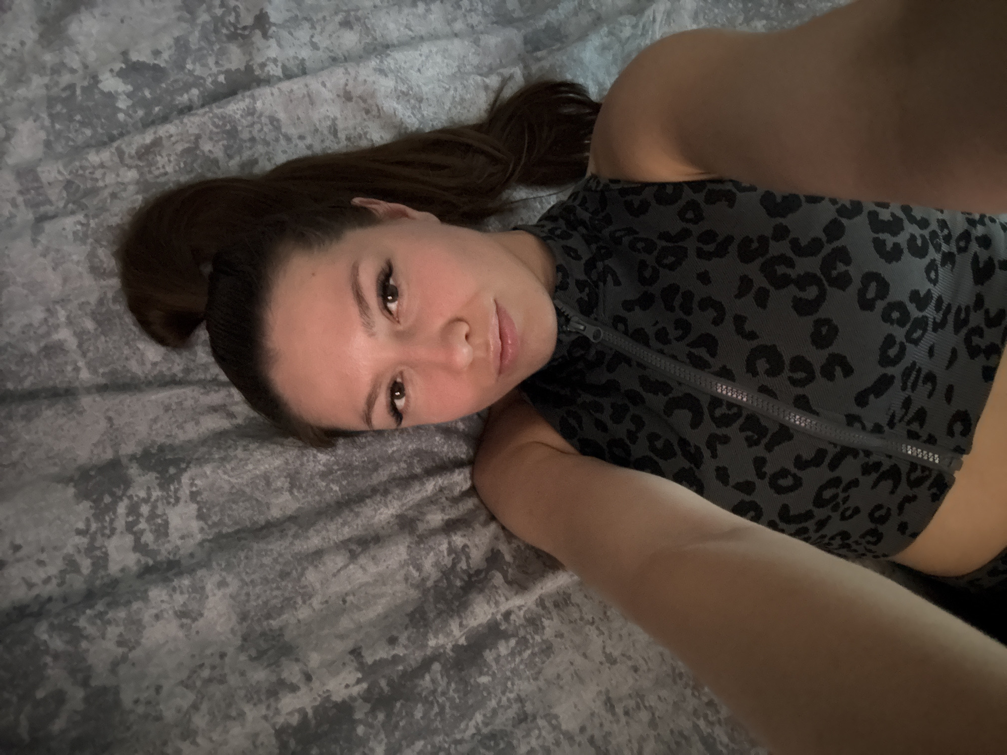 A person is lying on a textured grey surface, possibly a bed or couch. They are facing the camera with their head slightly tilted and a gentle smile. Their hair is dark brown and pulled back into a ponytail, with some strands falling across their face and shoulder. They are wearing a dark grey top with a black leopard print pattern and a zippered neckline. The lighting suggests an indoor setting, possibly during the day or evening. The overall mood conveyed is relaxed and natural. There is no visible text in the image.