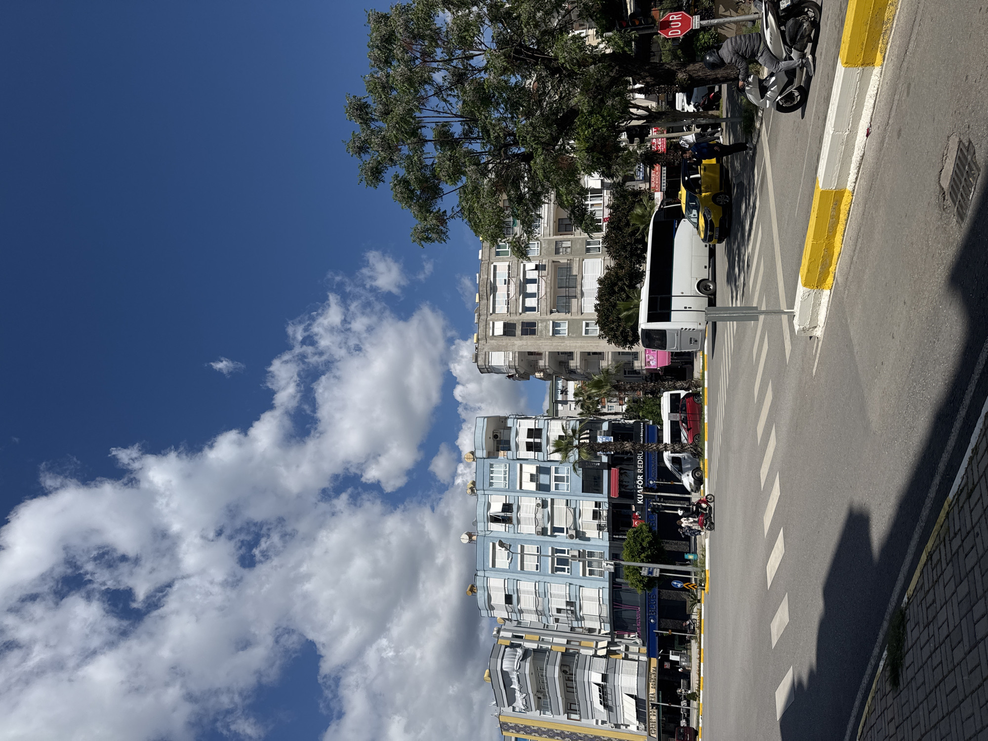 The image shows a street scene in an unidentified city. The sky is blue with scattered cumulus clouds. Buildings line the street, varying in height and architectural style. A large tree is present, its branches extending over the street. A pedestrian crossing is marked with white stripes. Vehicles, including a white bus, a yellow taxi, and several motorcycles, are visible. A stop sign is also present. People are standing or walking on the sidewalk. The street is paved, and the curb is painted yellow and white. The overall environment appears to be urban and sunny.