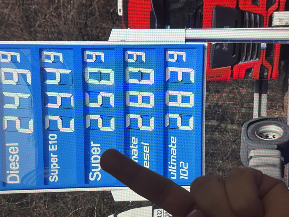 A close-up, slightly blurred image depicts a blue digital fuel price display at a gas station in Vellmar, Germany, observed during daylight hours. A light-skinned human hand with a visible index finger is positioned in the lower-left quadrant, pointing towards the "Super" fuel line displaying a price of 2.50⁹.
The digital sign, rendered in white text and segment numbers against a blue background, lists the following fuel types and prices in Euros per liter:
- Diesel: 2.49⁹
- Super E10: 2.44⁹
- Super: 2.50⁹
- Partially obscured text, likely "Ultimate Diesel" or similar, with a price of 2.80⁹.
- Ultimate 102: 2.83⁹
In the background, out of focus, a substantial portion of a red semi-truck is visible to the right of the sign, including its front grille, headlights, and a German license plate holder. Below the sign, a large, dark gray fender and a wheel, possibly belonging to the same or another heavy vehicle, are present. The far background shows defoliated trees or dense bushes, indicating an outdoor, possibly natural, setting beyond the gas station.