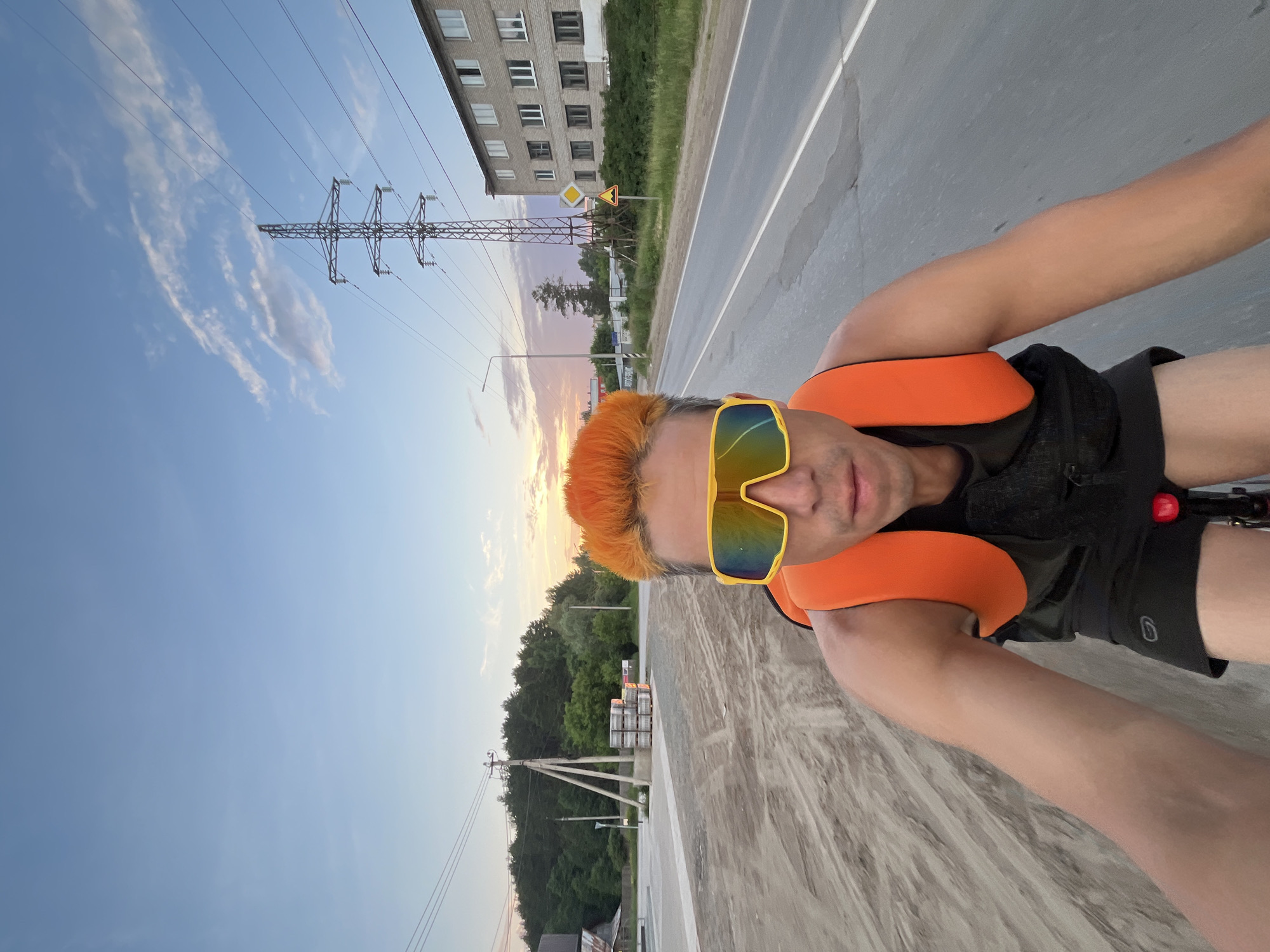 A person with bright orange hair and large yellow-tinted sunglasses is riding a bicycle on a paved road. They are wearing an orange vest over a black tank top and black shorts. The person is looking directly at the camera, holding it out to capture a selfie. The background shows a street with a building on the right side, a power line tower, and trees in the distance. On the left side of the road, there is a pile of construction materials and a sandy area. The sky is a clear blue with some scattered clouds, and the sun appears to be setting, casting a warm glow on the scene. The location is identified as Chaikovsky, Russia.