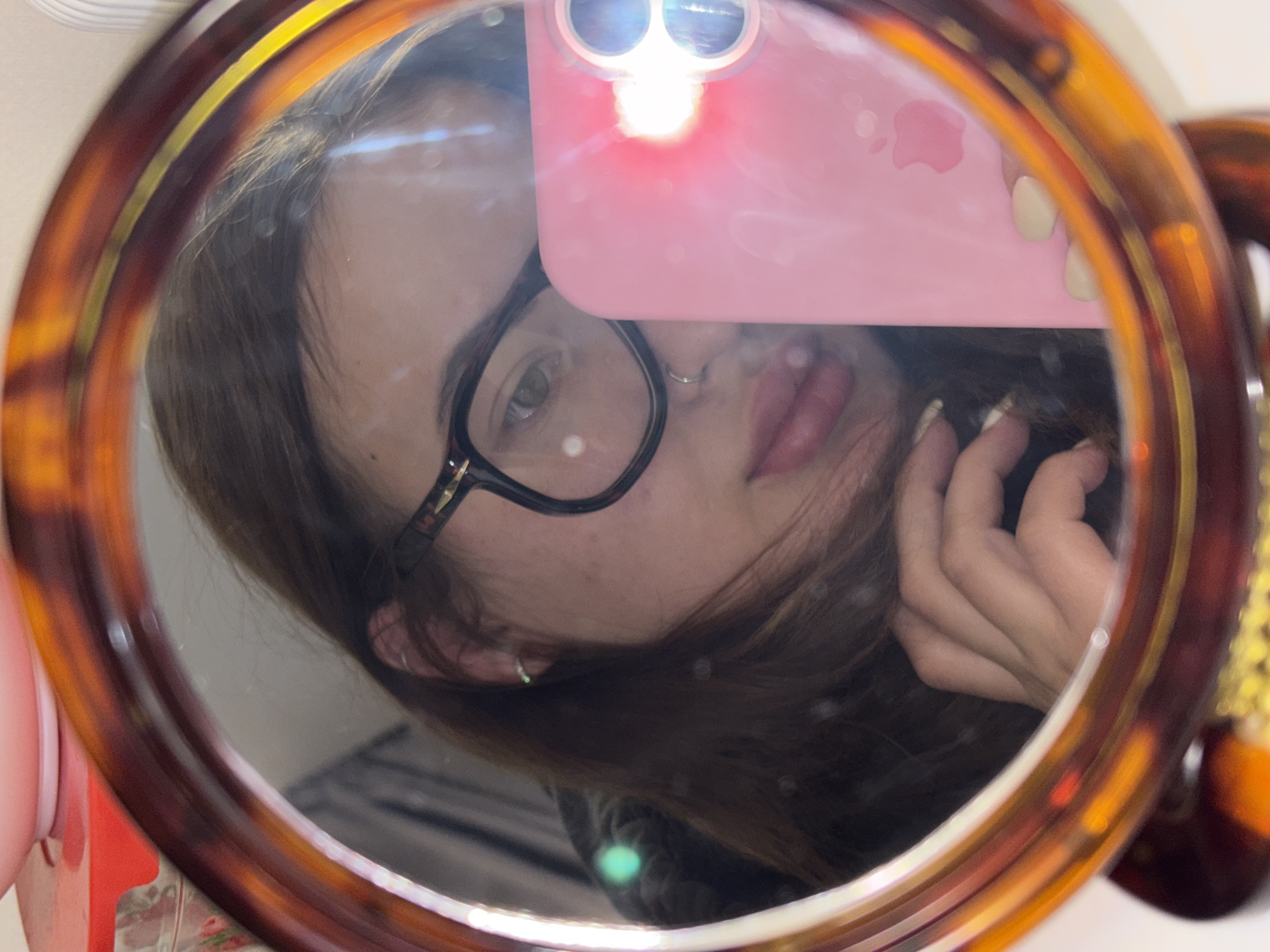 The image is a mirror selfie depicting a young woman, possibly taken in Krasnodar, Russia. She is reflected in a mirror with a distinct tortoiseshell-patterned frame that encircles the entire composition.
The subject is a young woman with long, dark brown hair that appears slightly tousled. She wears black-framed eyeglasses, through which her left eye is clearly visible, exhibiting a direct gaze towards the camera. Her lips are slightly parted, and a small silver hoop piercing adorns her right nostril. A subtle silver earring is visible in her left ear. Her fingernails are painted a light, possibly nude or cream, color.
The woman is holding a pink-cased smartphone, which is largely visible in the upper center of the reflection. The phone has two prominent rear camera lenses and emits a bright flash, creating a radiant highlight on her face and prominent lens flares on the mirror surface. The iconic Apple logo is clearly visible on the back of the pink phone case. The strong flash dominates the lighting, casting a bright illumination on her features and creating reflections across the mirror.
The overall scene is an informal, personal portrait captured through the act of taking a mirror selfie. The background reflected behind her is indistinct due to the close-up nature of the shot and the mirror's surface.