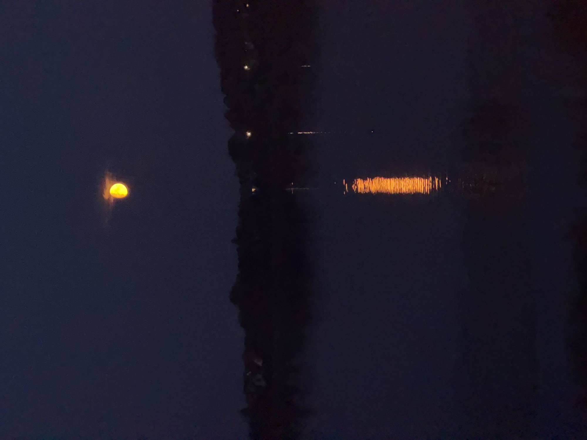 The image depicts a nighttime scene over a body of water, likely a river or lake. The sky is a deep, dark blue. A bright, orange-yellow, crescent-shaped moon is visible in the upper portion of the sky, partially obscured by a wispy cloud. Below the moon, a silhouetted treeline stretches across the horizon, with faint lights suggesting buildings or houses nestled within the trees. The water in the foreground is dark and reflects the moonlight and the lights from the shore, creating shimmering streaks of light. A faint mist hovers over the surface of the water. The overall impression is one of tranquility and natural beauty. There are no people or specific identifiable objects within the scene that would allow for precise location identification in Riga, Latvia.