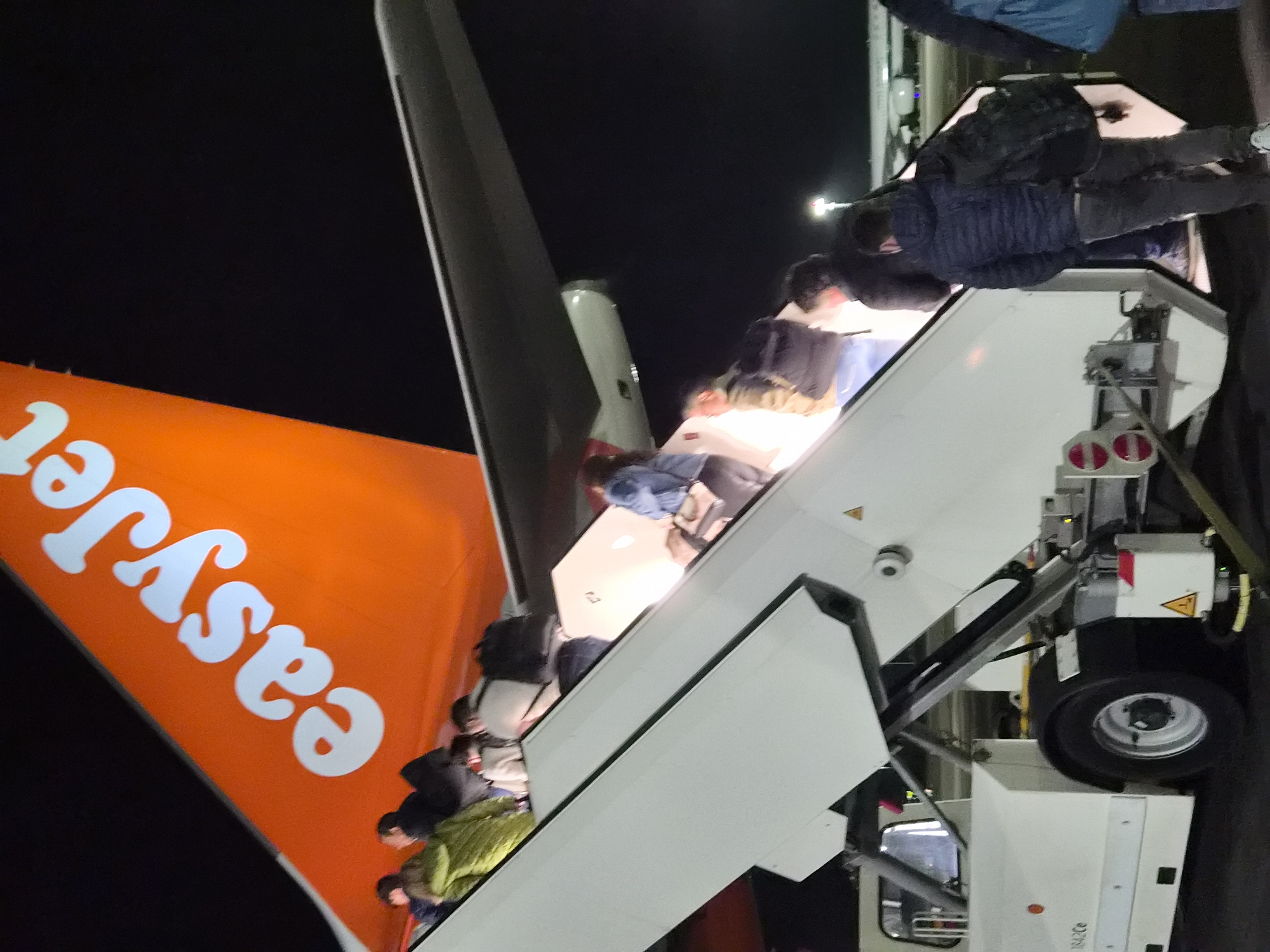 Passengers are disembarking from an EasyJet plane at night. The plane's tail fin, displaying the EasyJet logo in white lettering against an orange background, is visible. A mobile stairway is positioned against the plane's exit door, with a number of individuals ascending or descending the stairs. The city of Niederanven, Luxembourg, cannot be determined from the image.