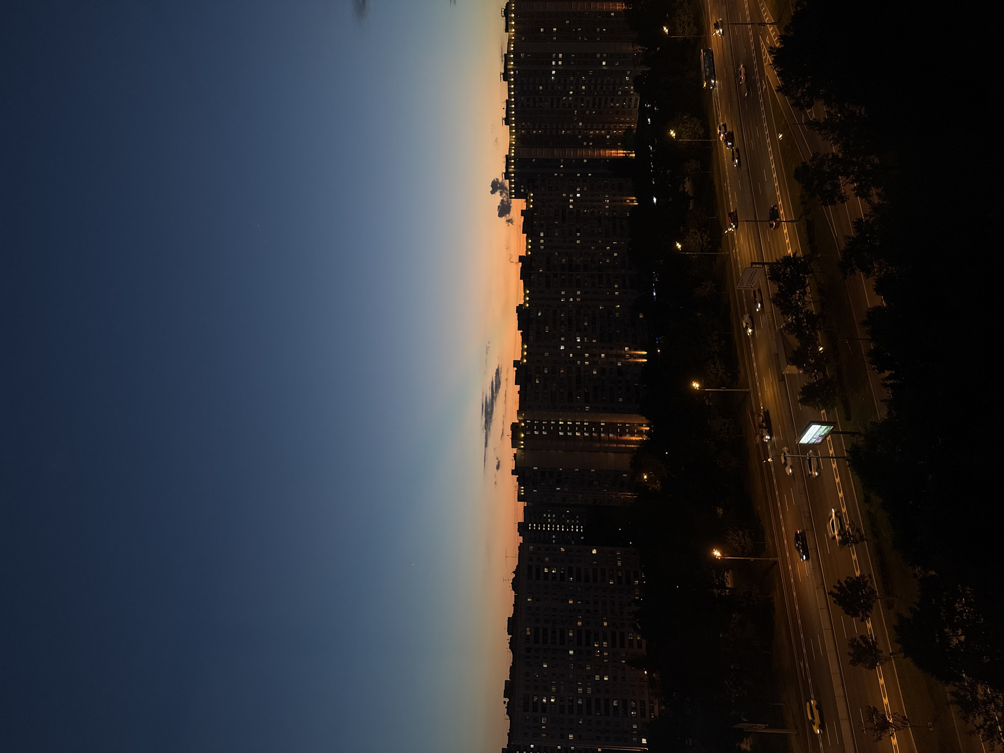 A wide shot captures an urban cityscape at dusk. The sky transitions from a deep blue at the top to a vibrant orange band near the horizon. Silhouetted against the fading light are numerous apartment buildings, their windows illuminated with warm light. In the foreground, a multi-lane highway is visible, with cars and a bus driving along it, their headlights and taillights creating streaks of light. Streetlights illuminate the roadway, and dark trees line the highway and obscure the lower portion of the scene. The overall impression is of a bustling city winding down for the night.