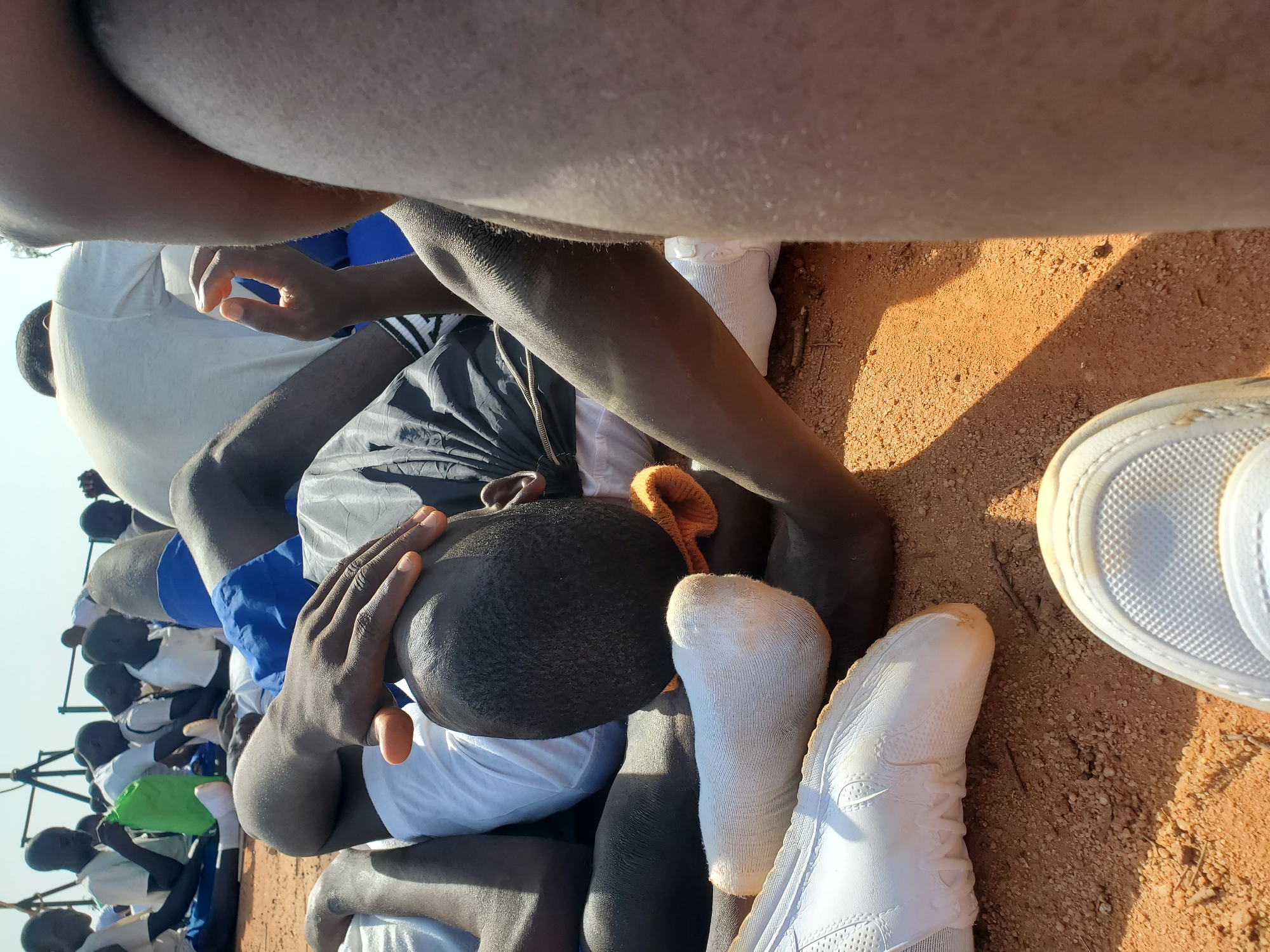 In Barakin Rafin Gora, Nigeria, a group of young men are gathered outdoors on a reddish-brown dirt surface, likely a field. The central focus is on one individual lying down, his head resting on the ground, with his hand placed on his forehead. He is wearing a white t-shirt and blue shorts. Another person's legs and arm are visible in the foreground, obscuring part of the central figure. Several other individuals are seated and lying down in the background, some wearing white shirts and blue shorts, and one is holding a green bag. The environment appears to be a sunny day, with a shadow cast across the ground. The scene suggests a break or rest period for the group.