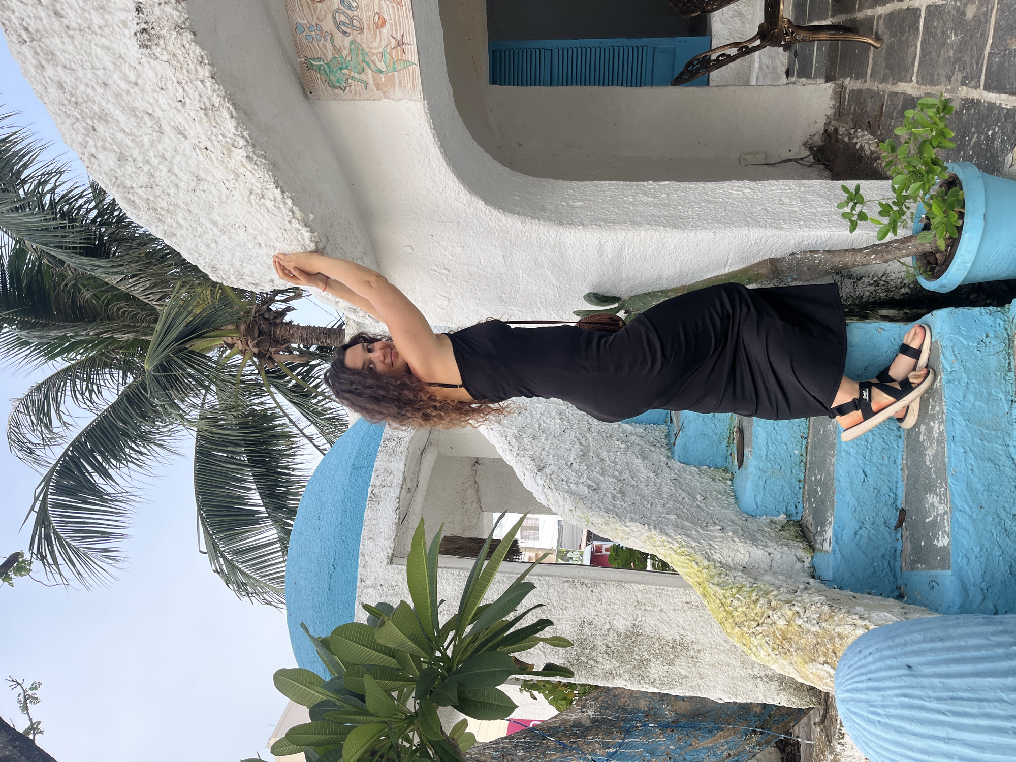 A young woman with long, curly brown hair is leaning against a white, textured wall with blue accents. She is wearing a black sleeveless dress and black sandals with white soles. Her arms are stretched above her head, and she is smiling at the camera. The scene appears to be outdoors, with lush green palm trees and other foliage visible. A bright blue planter with a small tree is situated on a step to the right of the woman. The architectural style is unique, featuring curved white structures and vibrant blue painted steps. The overall ambiance suggests a warm and sunny day, possibly in a tropical or subtropical location like Phu Thuy Ward, Vietnam.