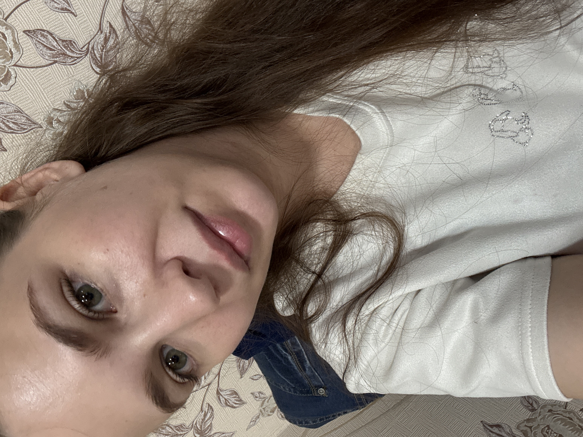A close-up, overhead shot shows a young woman lying down, her head tilted to the side. She has long, brown hair that cascades over her face and shoulders. Her skin is fair and appears smooth, with some light makeup on her eyes and lips, which are slightly parted. Her eyes are open and green, looking directly into the camera. She is wearing a white t-shirt with subtle silver embellishments that resemble cartoon characters. Beneath the t-shirt, a hint of blue denim is visible, suggesting she might be wearing overalls or a denim jacket.
The background is a patterned fabric, likely a couch or bedspread, with a floral design in muted beige and brown tones. The lighting appears to be indoor, casting soft shadows. The image conveys a relaxed and casual mood. No text is visible in the image. The location context provided is Nizhnyaya Gar', Russia, suggesting this is a personal, indoor photograph taken within that region.