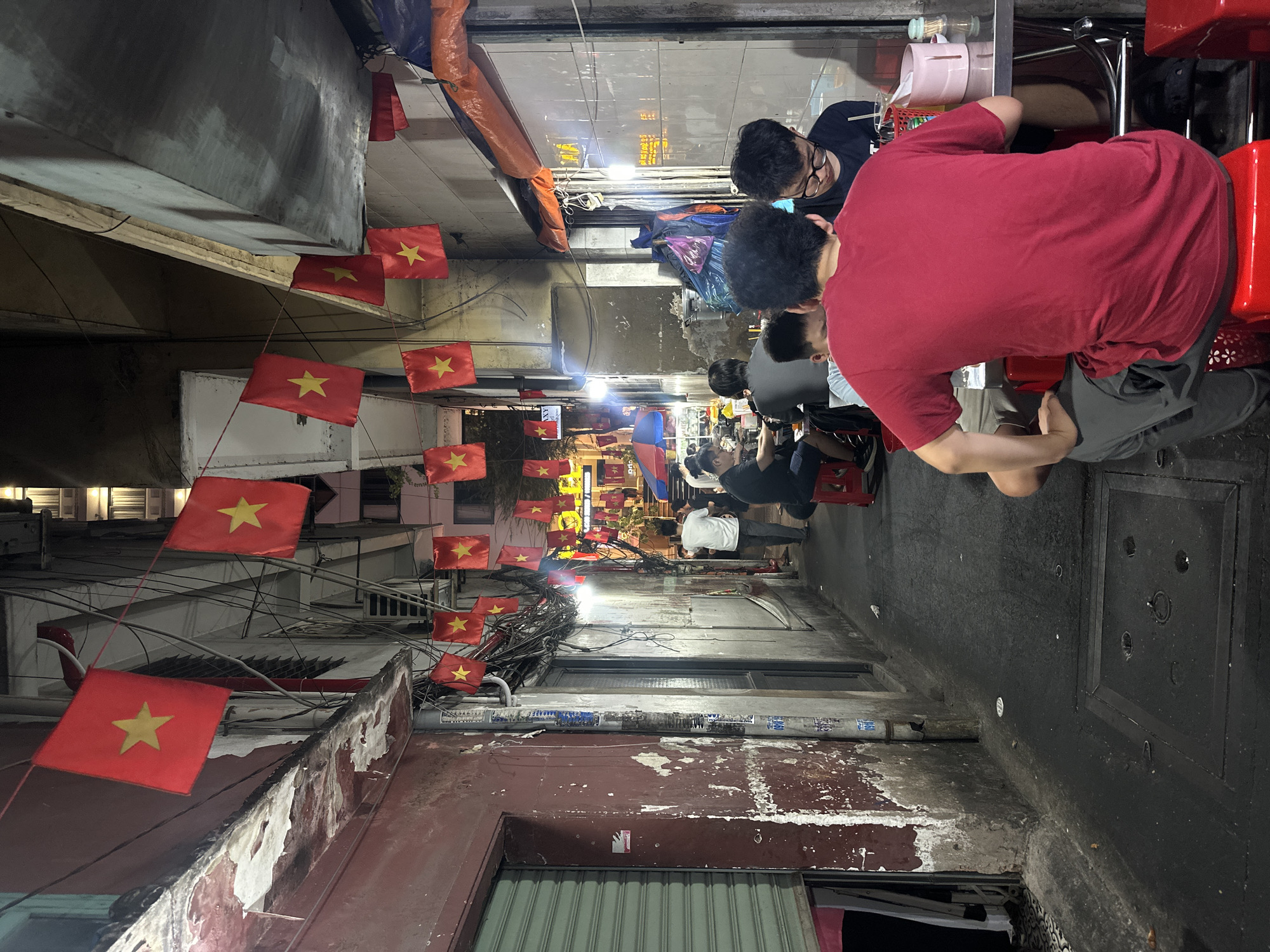 A narrow alley in Thủ Đức, Vietnam, is adorned with Vietnamese flags strung across its width. People are seated on small stools, presumably eating, with a food stall visible in the background. The alley is dimly lit, and the buildings on either side show signs of age and wear. Overhead wires and cables are intertwined, adding to the cluttered feel of the scene.