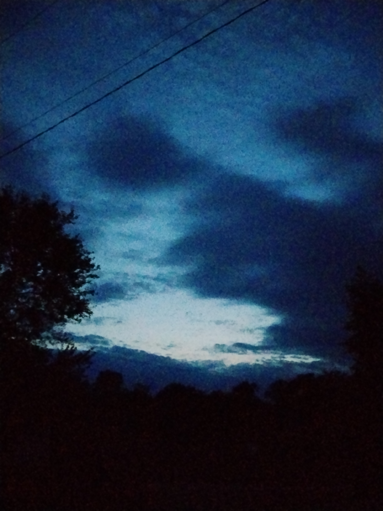 The image depicts a dark, moody sky with scattered clouds over a silhouetted landscape. A thin power line is visible diagonally across the upper portion of the frame. The lower half of the image is dominated by the dark shapes of trees and foliage, suggesting an outdoor setting at dusk or after dark. The sky is a deep blue, with lighter patches of clouds illuminated by an unseen light source, possibly the moon or a distant city light. The overall impression is one of quiet solitude and a natural environment. The image does not provide any specific indicators of the city of Ustyvytsia, Ukraine.