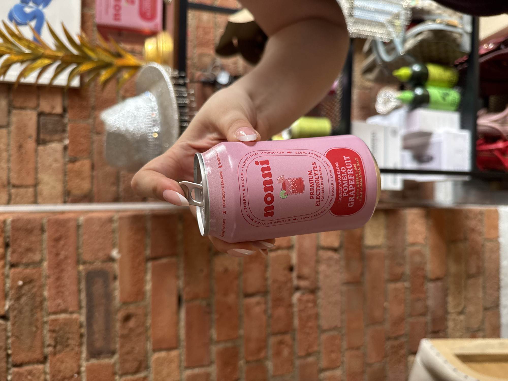 A person's hand is holding a pink can of "nonni" brand premium electrolytes, lightly sparkling pomelo grapefruit flavor, in front of a brick wall. The can is partially open, with the tab lifted. In the background, a sequined hat sits on a shelf next to a yellow decorative plant. Another pink box with the "nonni" logo is visible further back. The overall setting appears to be indoors, possibly a store or home, with shelves filled with various items.