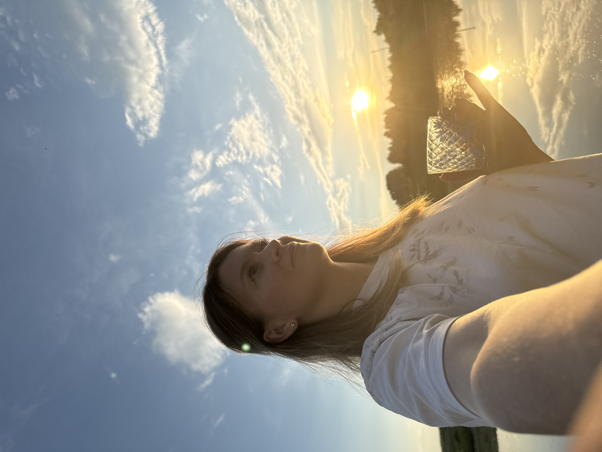 A young woman stands outdoors, holding a glass, as she looks towards the setting sun. The woman has blonde hair and is wearing a white t-shirt with a pattern. She is positioned to the left of the frame, with the sun on the right. The background shows a body of water with reflections of the sky and trees. The sky is blue with scattered clouds, and the sun is low on the horizon, casting a warm glow. The image appears to have been taken during a sunset or sunrise.