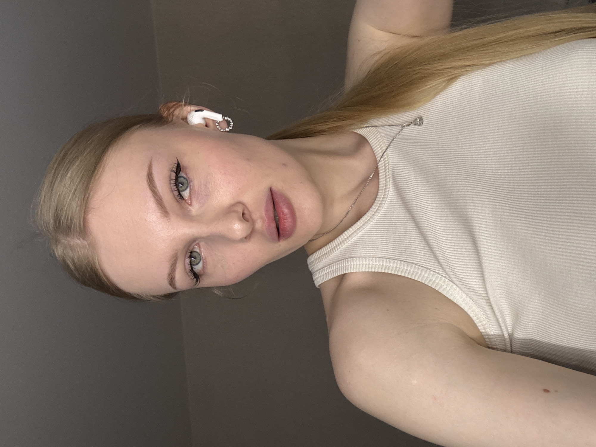 A young woman with light blonde hair pulled back into a ponytail is depicted in a close-up selfie. She is wearing a light-colored ribbed tank top and a silver necklace with a small pendant. A white earbud is visible in her left ear, adorned with a small, jeweled charm. Her eyes are light blue-gray, and she is wearing subtle makeup. The background is a neutral gray wall. There is no indication of Perm, Russia in the image.