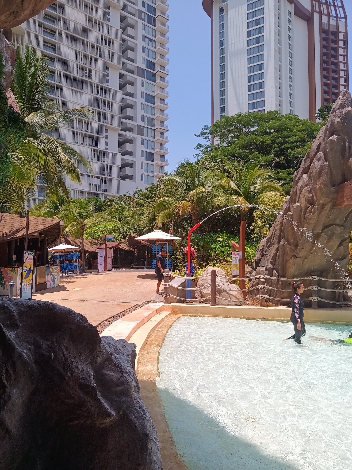 The photograph captures a daytime scene at a water park in Hua Hin City Municipality, Thailand. In the foreground, a shallow pool of water occupies a significant portion of the frame, with a person in a long-sleeved wetsuit walking in the water. To the left of the pool is a large rock formation, partially obscuring the view. Behind the pool, a path leads to several small buildings with thatched roofs and white umbrellas, suggesting a commercial area within the park. A man in a black shirt and shorts is walking on the path near the water's edge. Water is being sprayed from a red fixture toward the pool. The background features two tall buildings, one with a white facade and multiple balconies, and the other a white high-rise with brown architectural details. Palm trees and lush greenery are scattered throughout the scene, adding to the tropical atmosphere. The sky is a clear, pale blue.