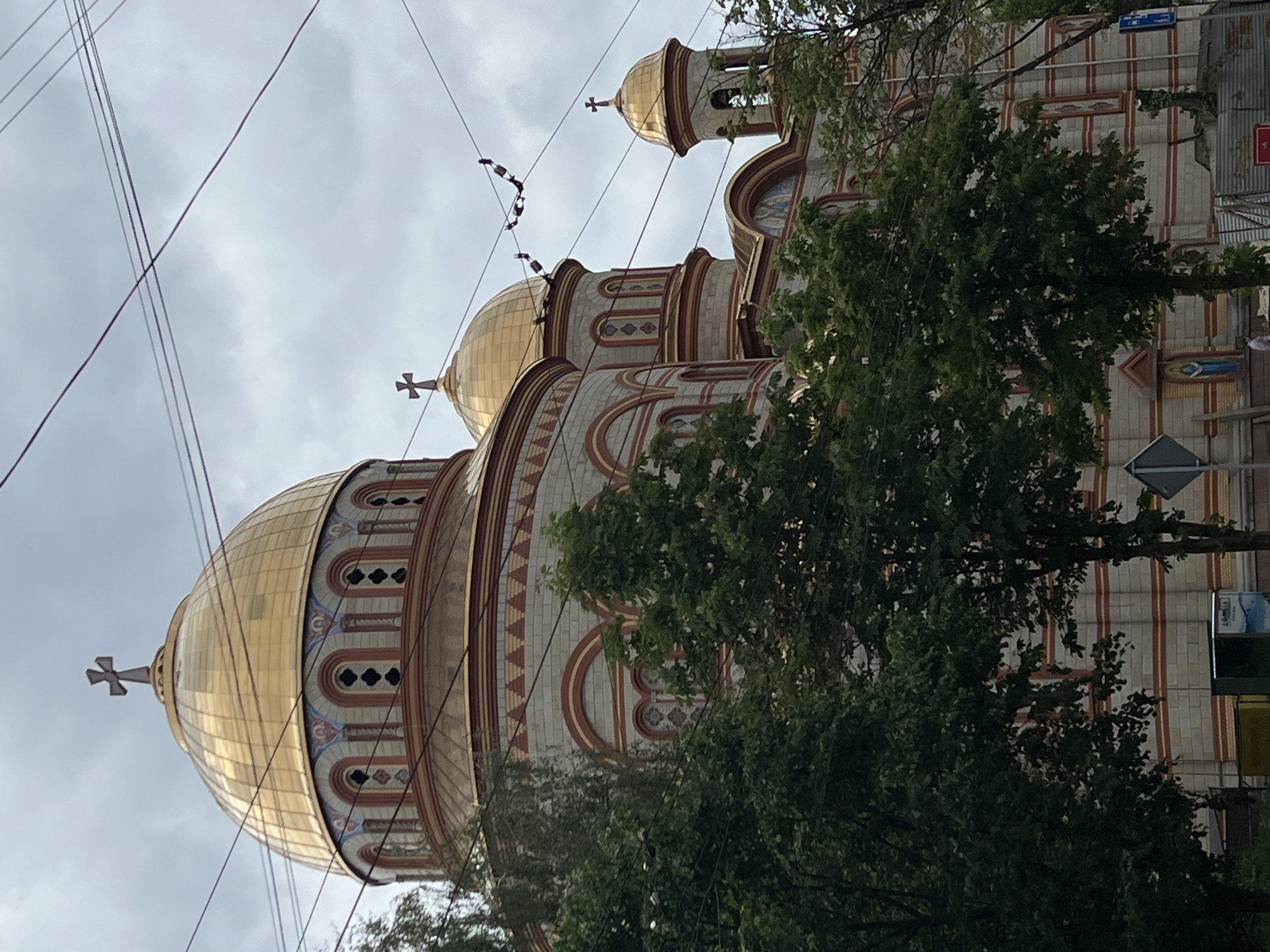 The image depicts the Nativity Church in Ternopil, Ukraine, from a low-angle perspective. The church is characterized by its golden domes topped with crosses. The building's facade features a mix of cream and red brickwork, adorned with arched windows and decorative patterns. A tree partially obscures the view of the church's base. Overhead power lines crisscross the sky. Street signs and a portion of adjacent buildings are visible to the right of the frame. The sky is overcast. There are no people visible.