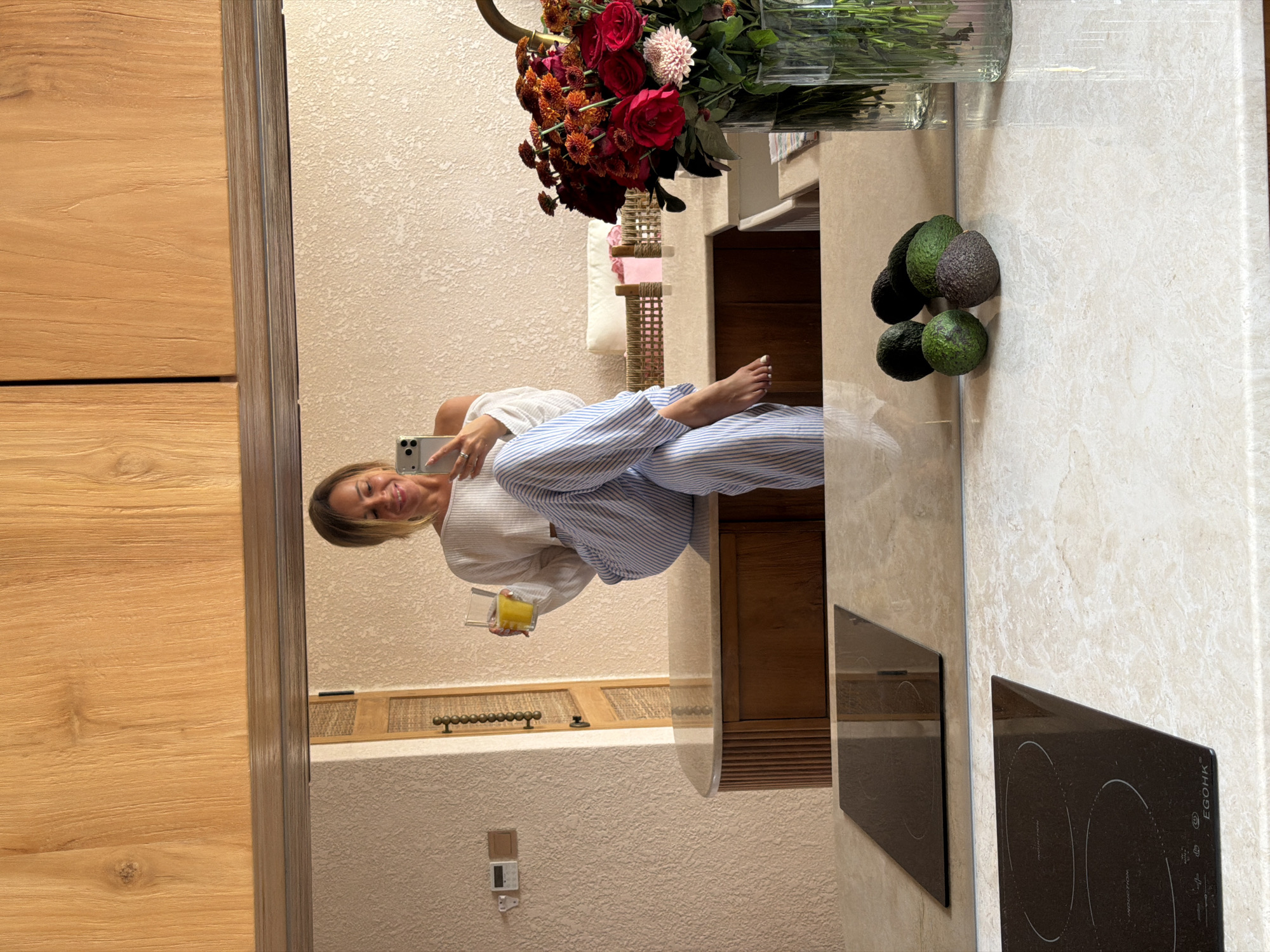 A mirror selfie reflects a woman holding a phone and a glass with a yellow liquid. She is wearing a white top and striped blue and white pajama pants. A vase with red and white flowers sits atop a countertop. Avocados are clustered near the vase. An induction cooktop is visible to the right of the woman’s reflection. The reflection reveals a textured wall. Wooden cabinets are visible in the reflection. The location is implied to be Mengwi, Indonesia, based on provided information.