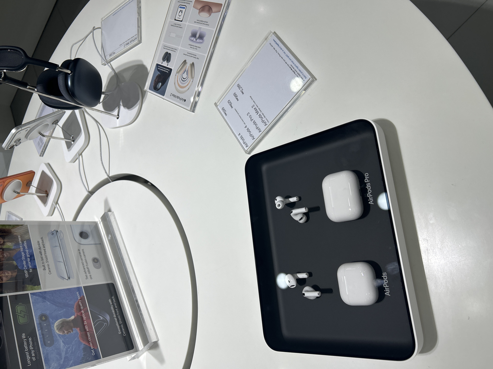 This appears to be a display of Apple products in a retail setting, likely an Apple Store, possibly in Kuala Lumpur, Malaysia, given the pricing tags in RM (Malaysian Ringgit). The products featured include AirPods (4th generation), AirPods Pro (possibly 3rd generation), and AirPods Max 2. There are several signage and displays with product information, including the price of the AirPods. There are also some iPhones shown in the display, highlighting features like camera and durability. The surfaces are white, and the lighting appears to be bright and even, consistent with a retail environment.