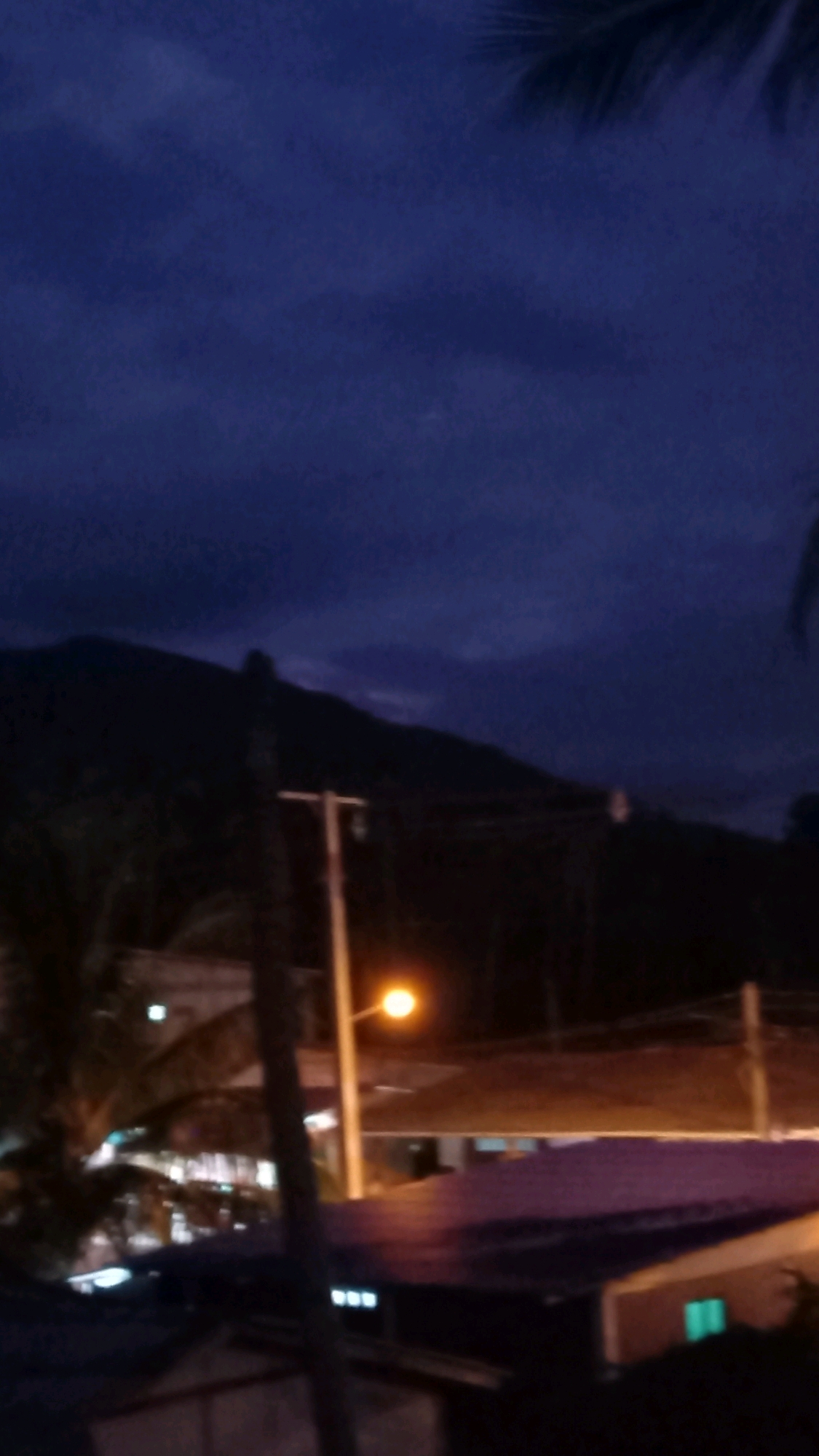 The image captures a dark, twilight scene with a prominent mountain range silhouetted against a deep blue, cloudy sky. In the foreground, several buildings are visible, some with illuminated windows and purple roofs. A streetlight with a bright orange glow illuminates a portion of the scene. A utility pole stands in the midground, and palm fronds are visible in the upper corners, suggesting a tropical location. The overall impression is one of a rural or semi-rural setting at dusk. The location is stated to be in the Ko Pha-ngan District, Thailand.
