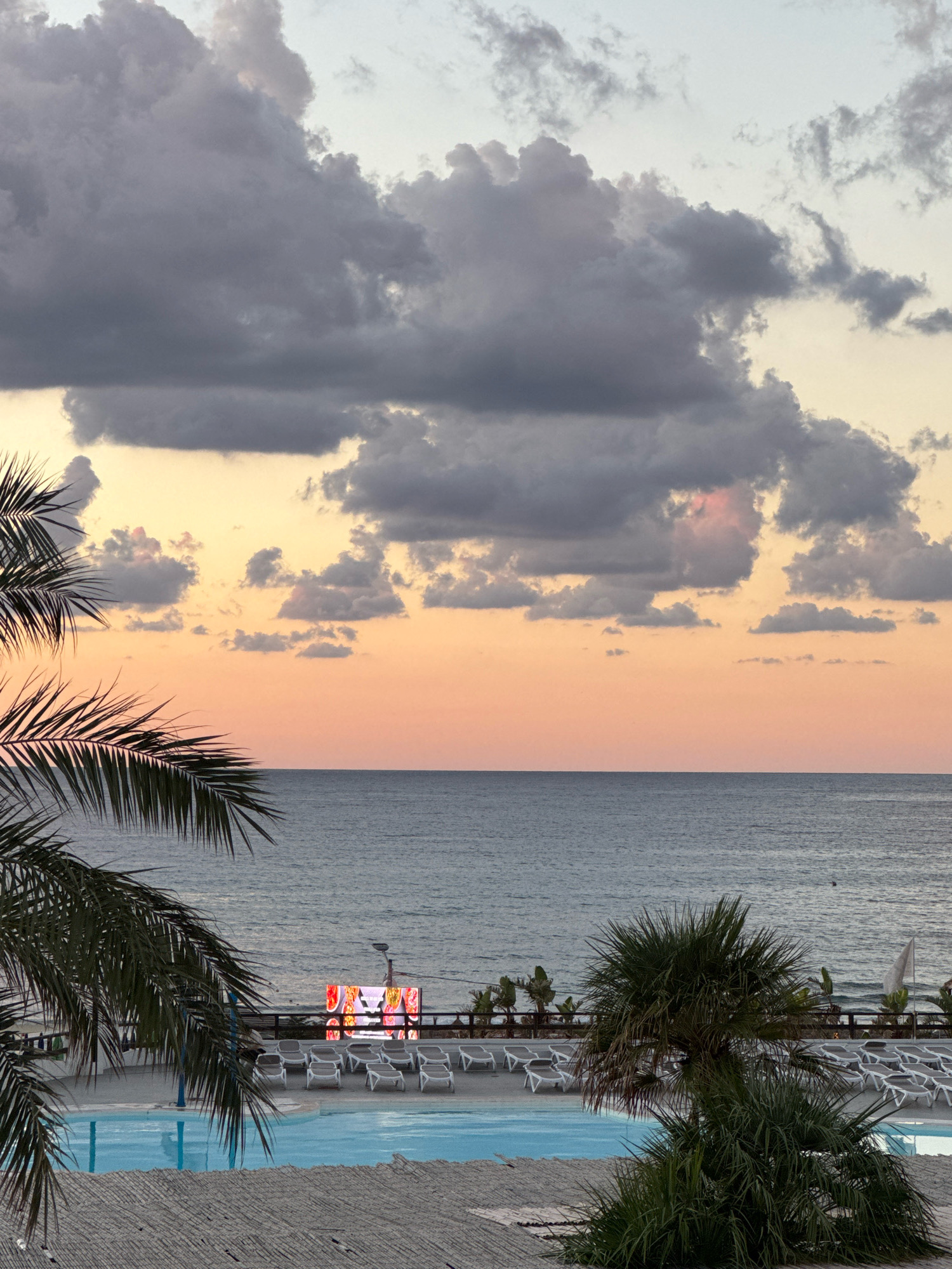 An outdoor scene in Capaci, Italy, features a resort area with a swimming pool in the foreground and the Mediterranean Sea in the background. The sky is filled with dark, dramatic clouds illuminated by an orange and yellow sunset. Palm trees frame the left and right sides of the image.
In the middle ground, a bright LED screen displays colorful images, likely advertisements or entertainment. Rows of empty lounge chairs are arranged around the pool, suggesting a beach resort or hotel. The water in the pool is a clear, bright blue, contrasting with the muted tones of the sea and sky. The environment appears tranquil, with no people visible in the immediate area.