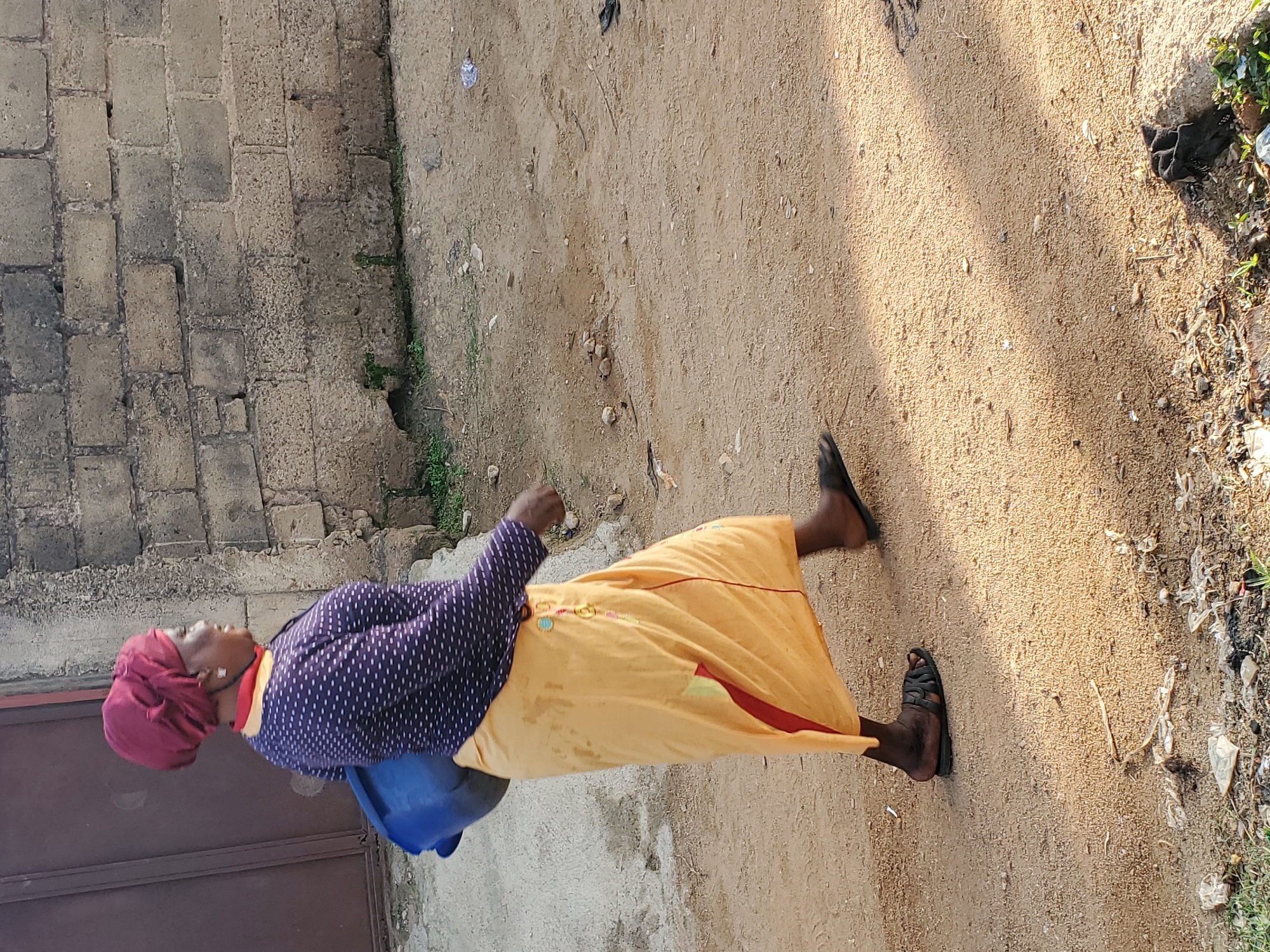 An older woman in Jos, Nigeria walks on a sandy path next to a weathered brick wall. She wears a burgundy head wrap, a dark blue, white-dotted blouse, and a long, pale yellow skirt. She carries a blue container. The path is littered with debris, and foliage grows on the other side.