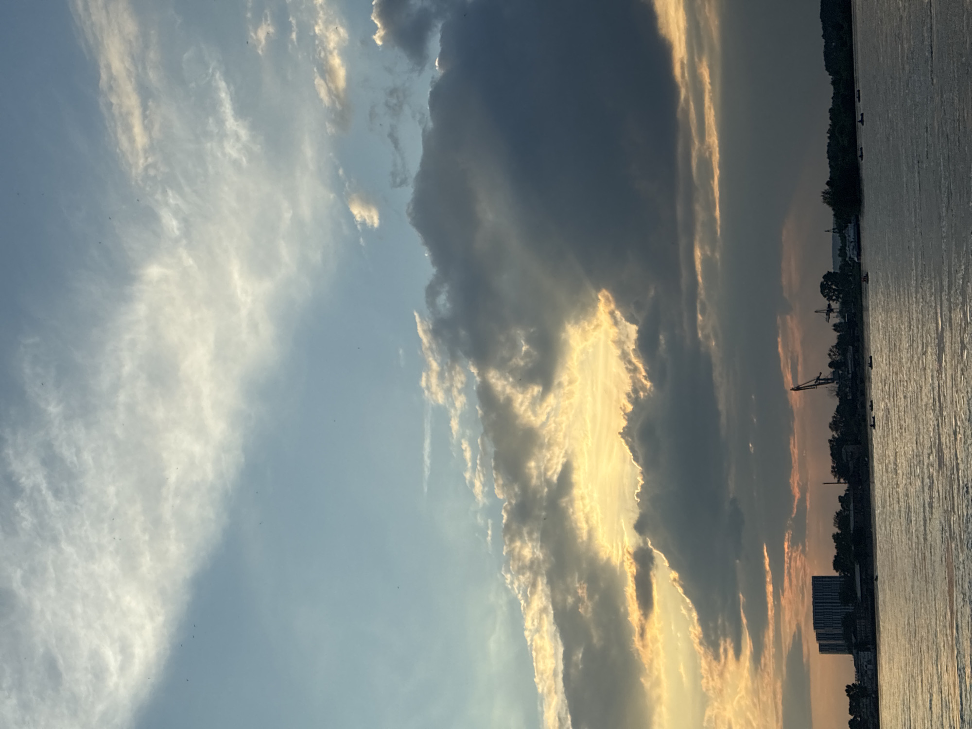 A wide-angle shot captures a hazy sunset over a body of water, possibly the Angara River, in Irkutsk, Russia. The sky is filled with dramatic clouds, some tinged with golden light from the setting sun, while others are dark and brooding. Silhouetted against the horizon are buildings, trees, and a large crane, hinting at an industrial or port area. Several small boats dot the water, suggesting recreational activity. The overall mood is tranquil, yet dramatic, due to the interplay of light and shadow in the sky.