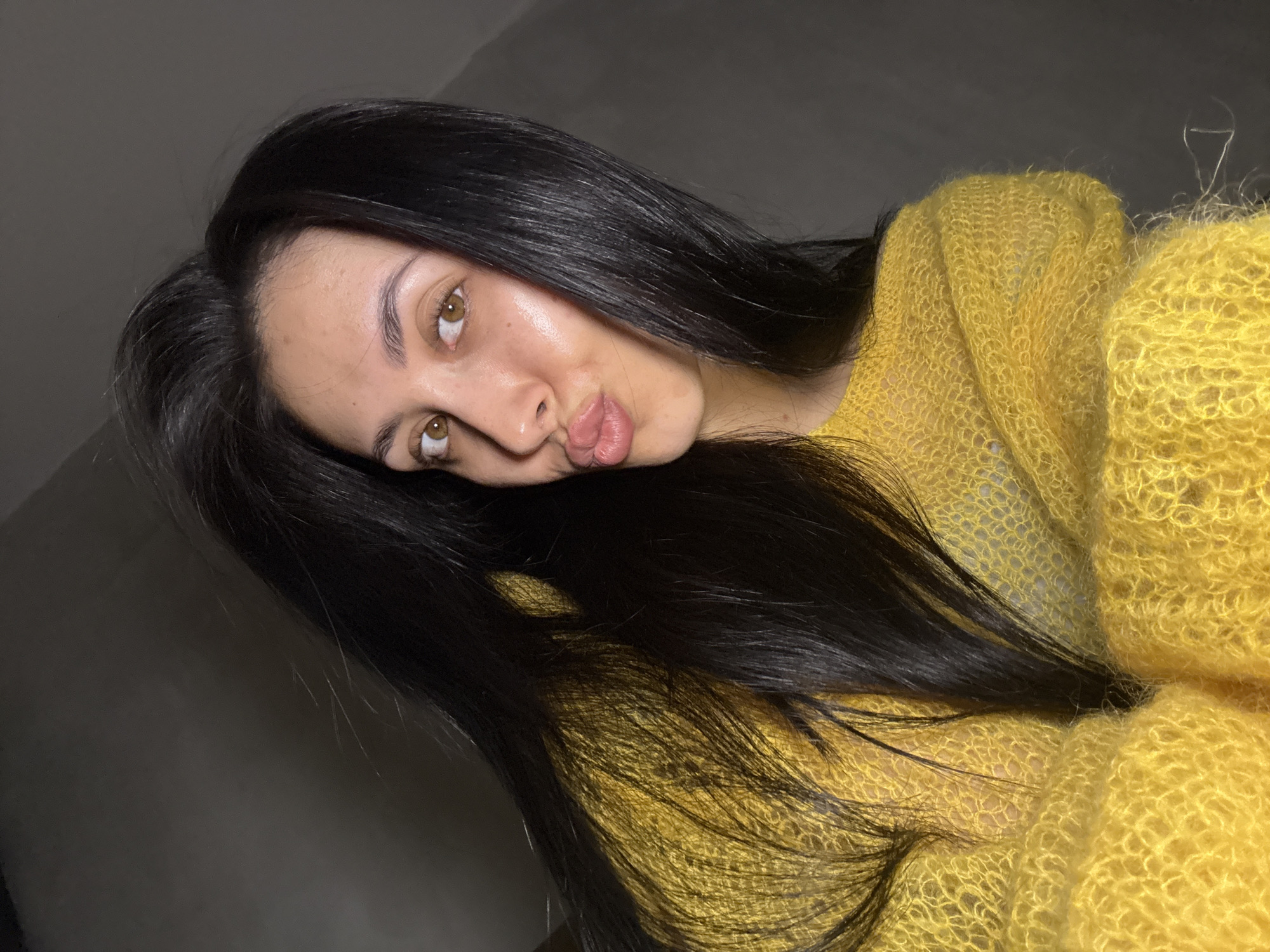 A young woman with long, dark hair is positioned at an angle in the frame, wearing a yellow fuzzy sweater. She is pursing her lips. Her skin tone is fair with noticeable moisture on her forehead and nose. The background is a dark, plain wall. The lighting is uneven, creating shadows. The location cannot be determined.