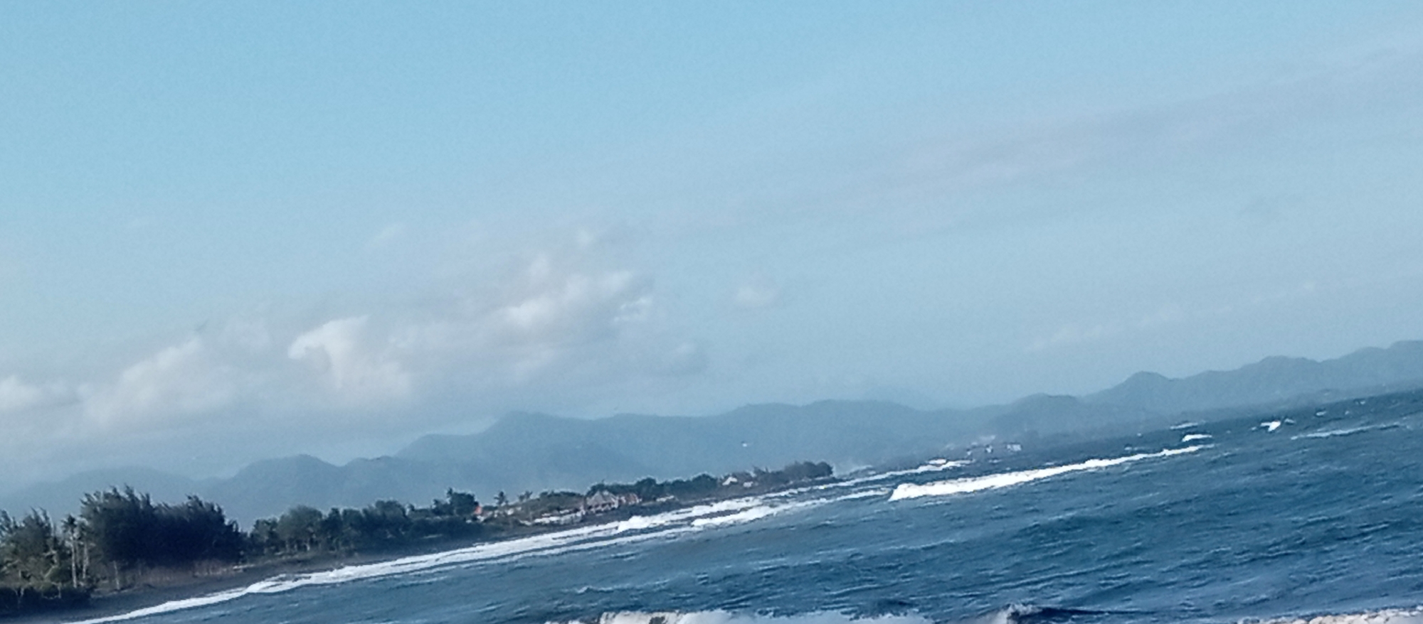 The image displays a coastline with turbulent ocean waves in the foreground. A stretch of dark sand beach is visible behind the waves, with a line of trees and a few buildings nestled amongst them. In the background, a hazy range of mountains stretches across the horizon under a pale blue sky with scattered clouds. The scene appears to be a natural landscape with some human development along the coast.