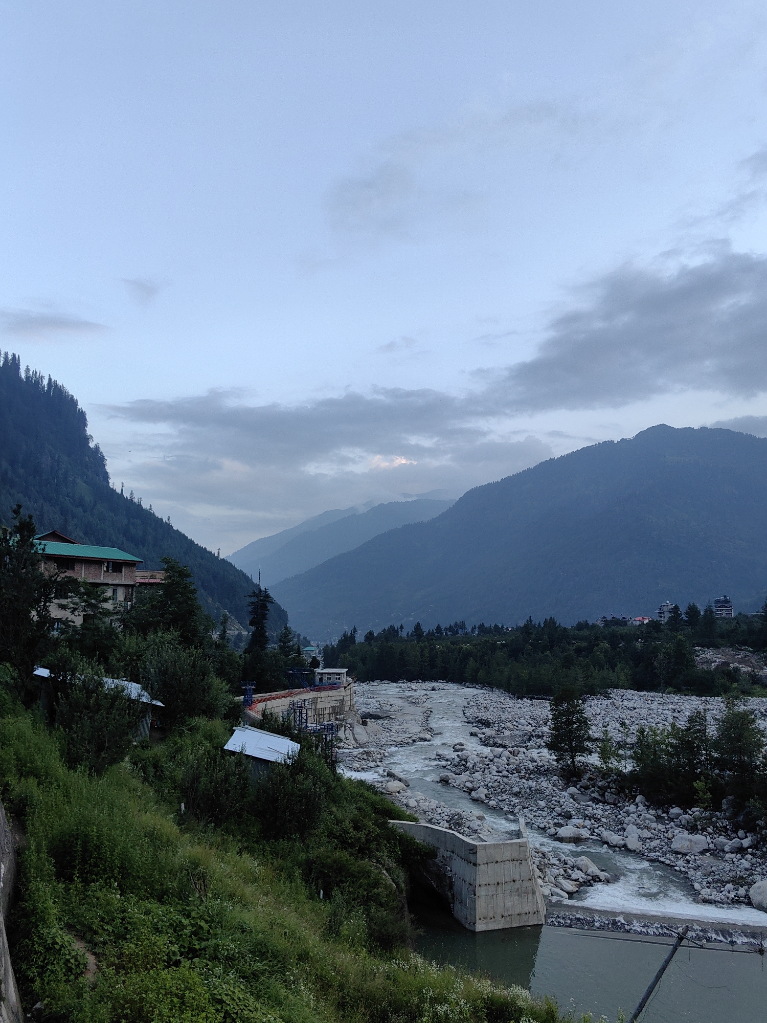 The image depicts a scenic view in the city of Kullu, India, characterized by a valley setting with rolling mountains and a river. The sky is overcast with grey clouds.
In the foreground, a steep, verdant hillside is covered in lush green vegetation and trees. Several small, metallic-roofed structures are nestled into the hillside, some appearing to be sheds or small dwellings. A concrete structure, possibly part of a dam or water management system, is visible at the edge of the hillside, leading down to the river.
The river itself flows through a rocky terrain, with numerous large grey stones lining its banks and bed. The water appears to be moving at a moderate pace, with some ripples and white water indicating flow. A concrete barrier or wall is built along one side of the river.
Further into the background, the valley opens up, revealing layers of mountains that recede into the distance. These mountains are densely covered with dark green coniferous trees, creating a textured, mountainous landscape. A few small buildings or clusters of houses are visible on the slopes and at the base of the mountains. A prominent building with a green roof is situated on the left side of the image, built on the mountainside.
The overall impression is one of a natural landscape with human settlements integrated into the mountainous environment. The lighting suggests it is either early morning or late evening due to the soft, diffused light and the subdued colors.