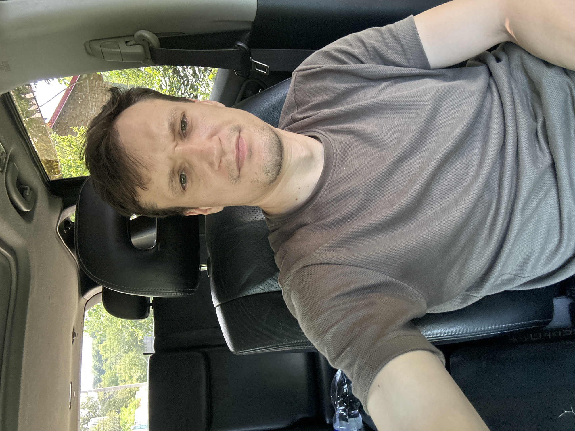 A man is sitting in the driver's seat of a car, facing the camera. The car appears to be parked outdoors, with trees and a building visible through the windows. The man is wearing a brown t-shirt and has short brown hair. He is looking directly at the camera with a neutral expression. The interior of the car is visible, including black leather seats and a sunroof. The image appears to be a selfie, taken from the perspective of the man. The location is the city of Makiv, Ukraine.