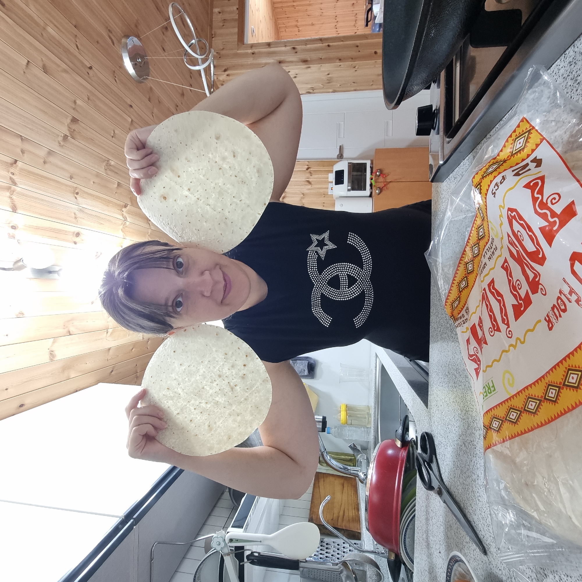 A woman stands in a kitchen, holding two large, round tortillas up to her ears. She is wearing a black t-shirt with a sparkling double-C logo. Behind her, a wooden ceiling with exposed beams is visible, along with a skylight and a modern light fixture. The kitchen counter is speckled gray, and to the left, a red pot sits in a sink filled with dishes. A pair of large scissors lies on the counter in front of a bag of tortillas. To the right, a stovetop with a cast-iron pan is visible, and a wooden cabinet with a white appliance sits against a wood-paneled wall. The image appears to have been taken indoors in a residential kitchen.