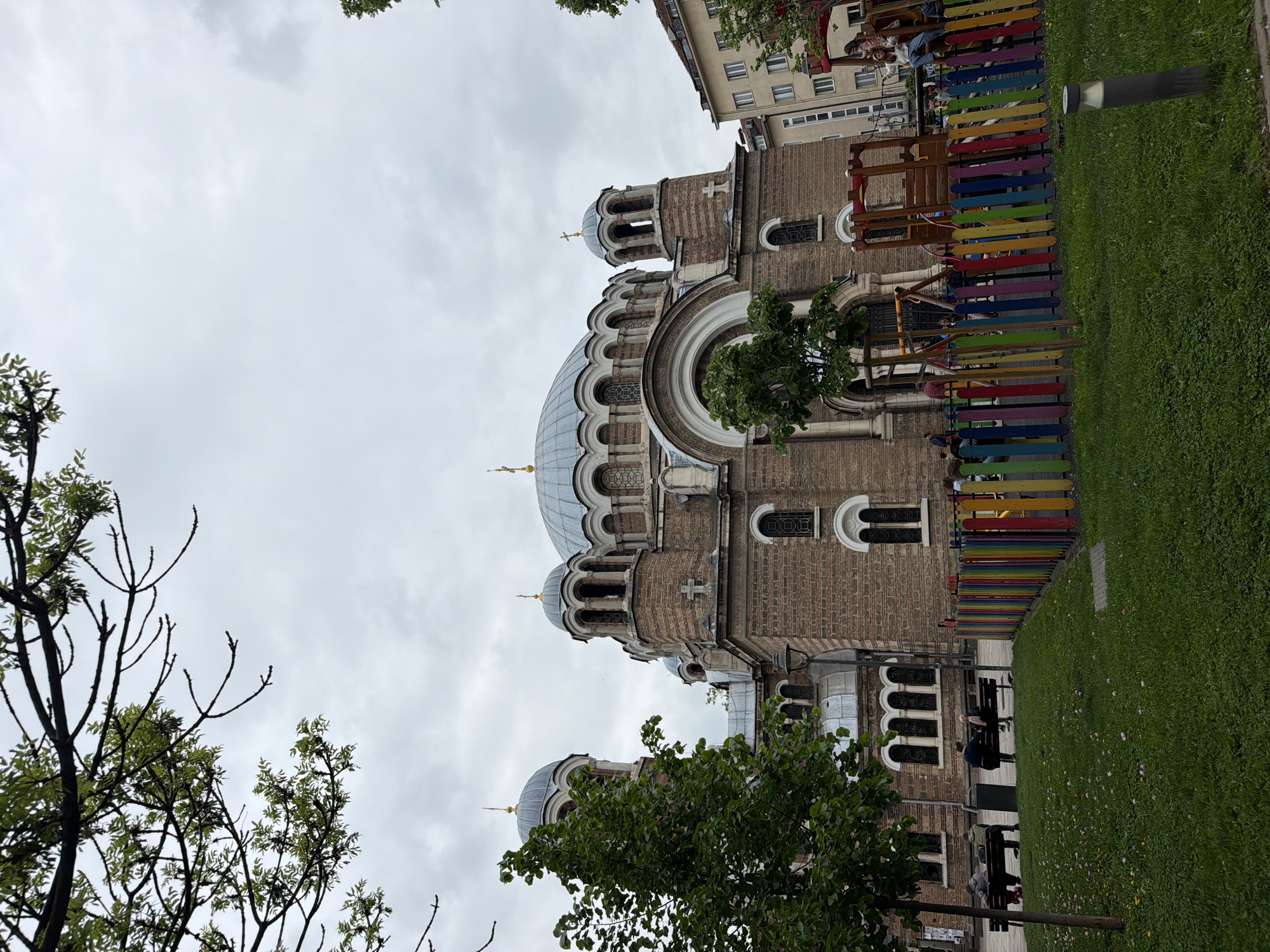 The image shows the St. Nedelya Church in Sofia, Bulgaria, shot from a slightly low angle, emphasizing its architectural details. The church, made of brick and stone, features a large central dome and smaller domes topped with golden crosses. Adjacent to the church, a playground with a colorful fence and equipment is visible. In the foreground, trees frame the scene. Some people sit on benches near the church. The sky is overcast.