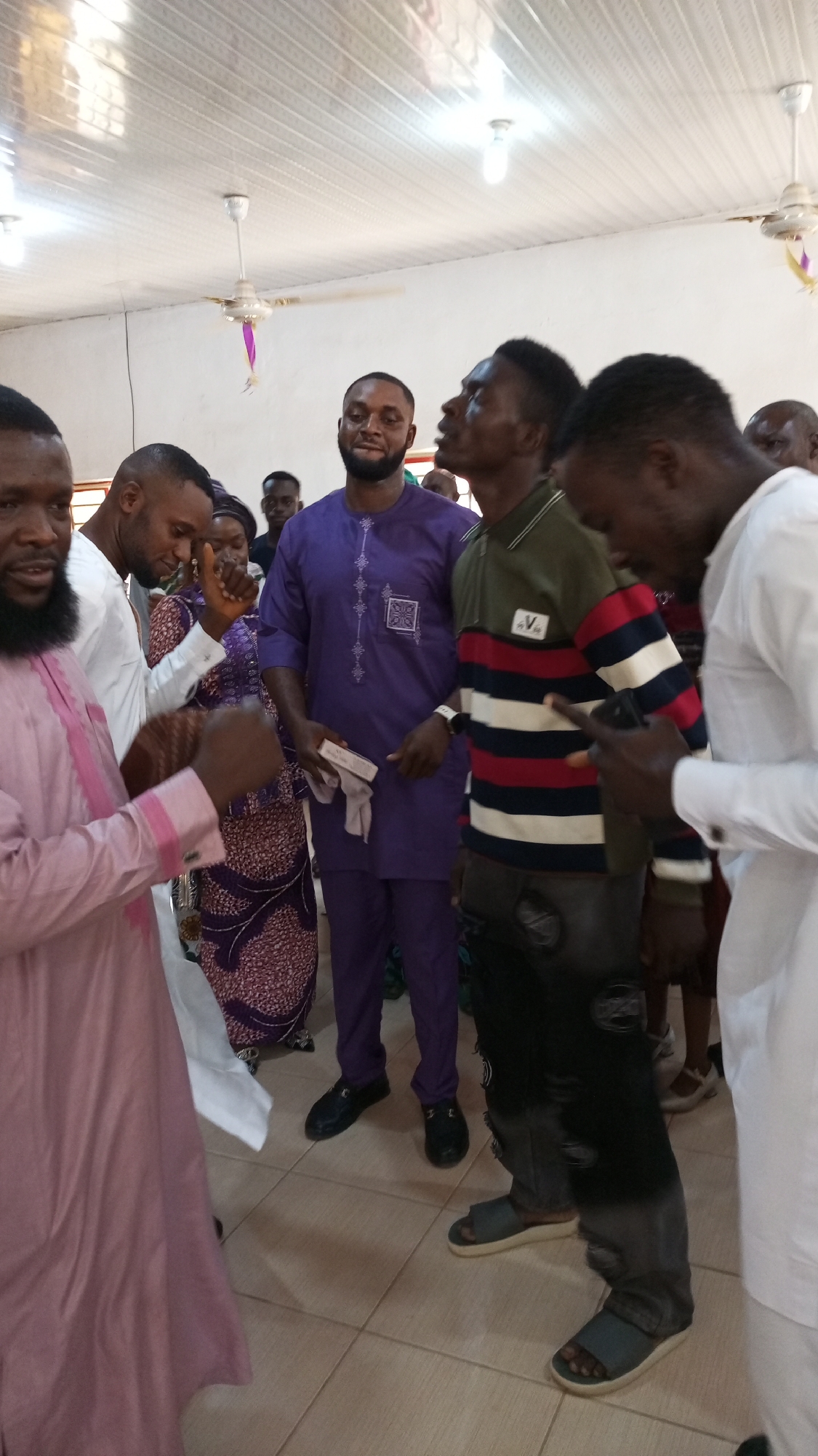 A group of people stand in what appears to be an indoor setting, possibly a meeting hall or church, given the bright overhead lighting and the presence of ceiling fans. The location is unspecified except for being somewhere in Sabon Tasha, Nigeria.
The individuals are dressed in a variety of clothing, including traditional African attire and more casual wear. There are men wearing robes in colors like pink, white, and purple, as well as a person in a striped sweater and jeans. The clothing styles suggest a mix of cultural and contemporary influences.
The people are generally standing close together, and some appear to be engaged in conversation or looking upwards. The environment is simple, with a tiled floor and white walls. The focus is on the individuals and their interactions, without a specific activity being clearly defined.