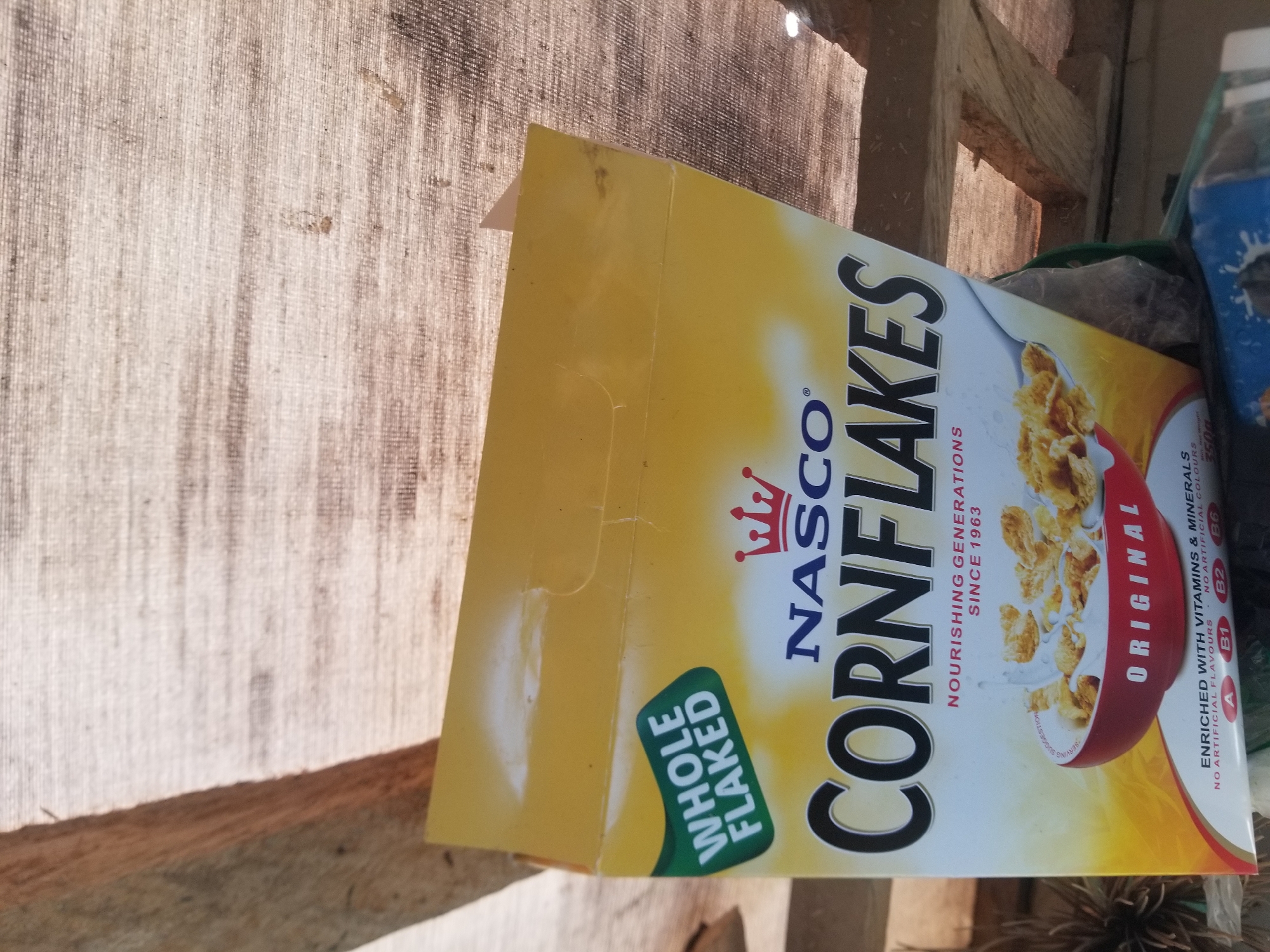 The image shows a partially obscured box of NASCO CORNFLAKES cereal, primarily its upper portion, against a backdrop of what appears to be a woven material and wooden beams. The cereal box is yellow and white, with prominent black text reading "CORNFLAKES". The brand name "NASCO" is displayed above "CORNFLAKES" with a crown logo. A green label on the left side reads "WHOLE FLAKED". Below the cereal name, it states "NOURISHING GENERATIONS SINCE 1963". A red bowl filled with cornflakes and milk is visible on the lower part of the box. Text at the very bottom indicates "ORIGINAL" and "ENRICHED WITH VITAMINS & MINERALS". The image seems to have been taken in an outdoor or storage setting with natural lighting. The specific location of Jauro Yinu, Nigeria, cannot be determined from the visual information provided in the image.