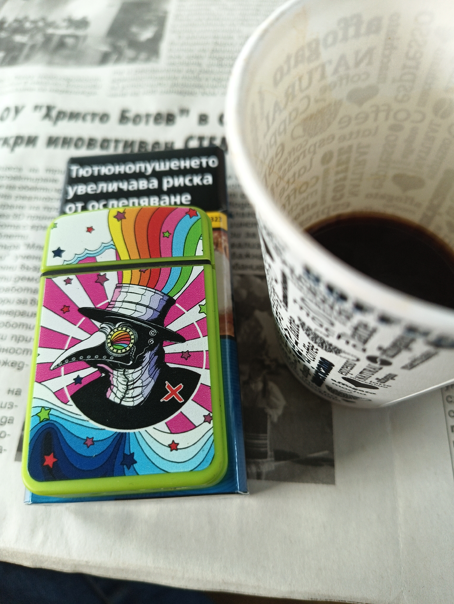 The image shows a close-up of several objects on top of a newspaper. The newspaper has text in Bulgarian, including "ОУ 'Христо Ботев' в" and "кри иноватИВЕН СТЕА". There is a brightly colored lighter with a cartoon image of a plague doctor wearing a black hat and cloak with a red "X" on the chest. The background of the lighter features stars, rainbows, and waves. Next to the lighter is a partially empty coffee cup with coffee-related words printed on its surface, such as "espresso" and "latte." The cup contains some dark coffee at the bottom. A cigarette pack is also partially visible beneath the lighter.