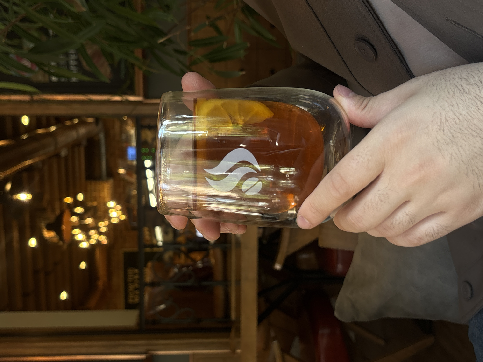 A person is holding a double-walled glass containing a brown liquid, possibly tea or a cocktail, with a slice of lemon inside. The glass features a white logo. The person is wearing a gray-brown suit jacket over a light shirt. The background appears to be the interior of a restaurant or cafe, with warm lighting, exposed wooden beams, and shelves with various items. The location is Yerevan, Armenia, as suggested by the lighting and decor. The person is holding the glass at eye level, possibly about to take a drink.