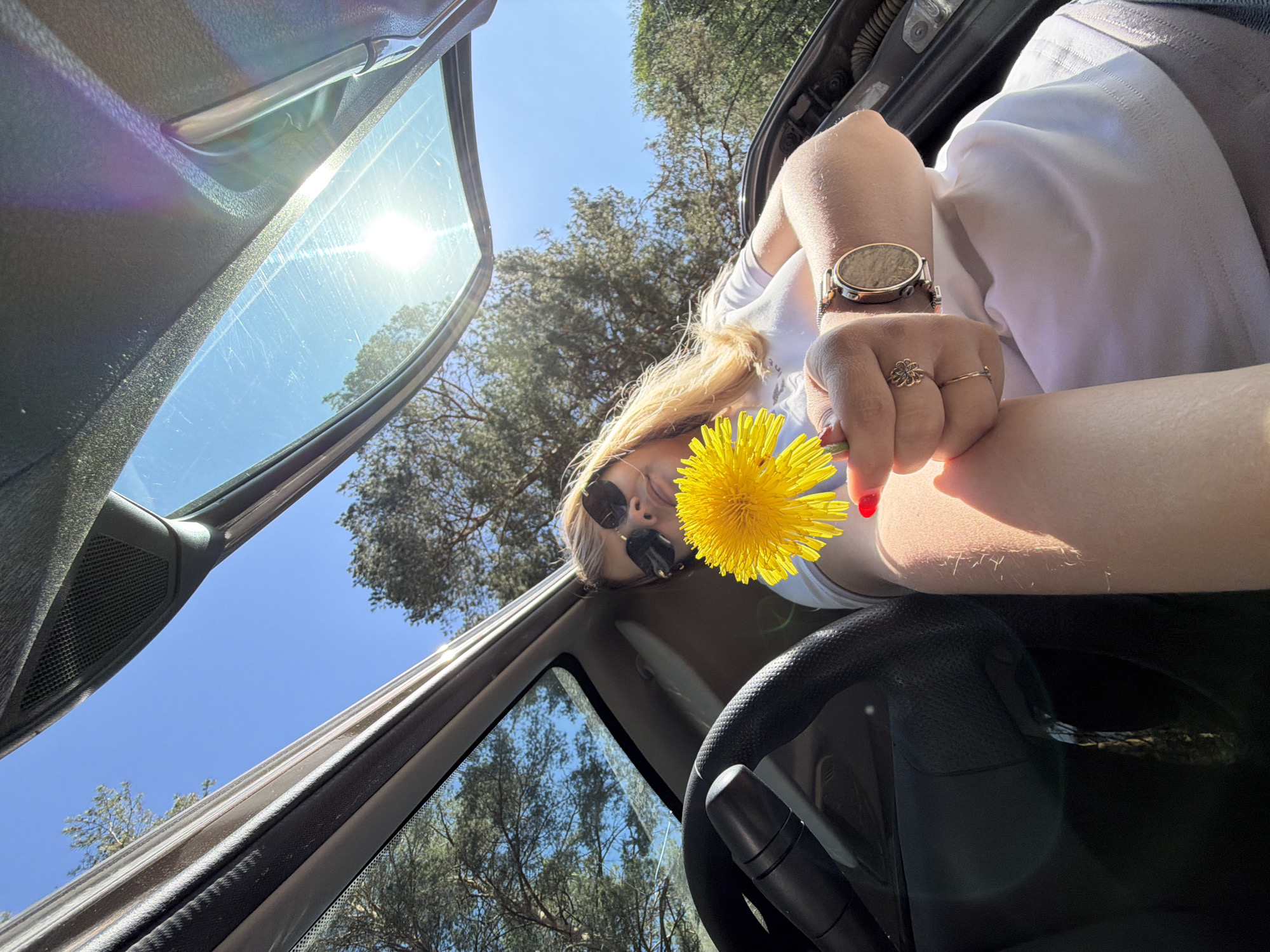A low-angle shot looking up into the interior of a car, revealing a fair-skinned person holding a yellow dandelion flower. The person is wearing sunglasses, a white shirt, and a wristwatch on their left wrist. Their fingernails on the hand holding the flower are painted red. They also have a ring with a flower design on their finger. The car door is open, and blue sky and trees are visible through the open space. The steering wheel is visible at the bottom of the image. The scene is bathed in sunlight. No information to determine the location.