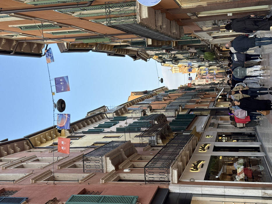 This image depicts a bustling pedestrian street in the city of Verona, Italy, likely Via Giuseppe Mazzini or a similar commercial thoroughfare, with the prominent Lamberti Tower visible in the distance. The scene is observed from a slightly elevated perspective looking down the length of the street.
The street is flanked by multi-story historical buildings, typically four to five floors tall, featuring facades painted in various warm hues including terracotta, yellow, and peach. Many windows are equipped with dark green louvered shutters, some open, some closed. Numerous ornate black wrought-iron balconies protrude from the upper floors. The ground level of these buildings is occupied by retail establishments, characterized by large display windows, some reflecting interior lighting. A visible shop on the left features illuminated signage that reads "Cialdea."
Overhead, a series of banners and a circular dark object are suspended across the street on wires. The banners display diverse graphics: one is orange with white Chinese characters and numbers "14 14"; another is blue with a stylized basketball design; a third blue banner also features white Chinese characters.
The paved street is densely populated with approximately twenty to thirty individuals walking in both directions. People are casually dressed, indicating a typical daytime or early evening scene. Several individuals are seen conversing or walking in small groups. Notable figures include a woman in a black dress carrying a brown handbag, a man in a dark jacket and jeans walking alongside her, and further down the street, individuals wearing hoodies with visible text such as "YUKUS" and "SWINE" (partially obscured). The general activity suggests pedestrian traffic for shopping or leisure. The sky above is clear blue.